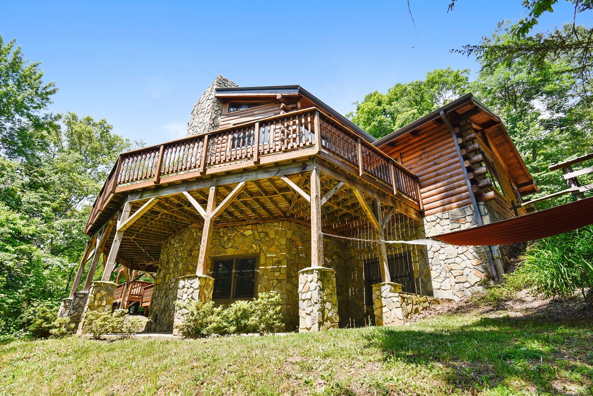 Stone and log cabin with a wooden deck, nestled in lush greenery under a blue sky.