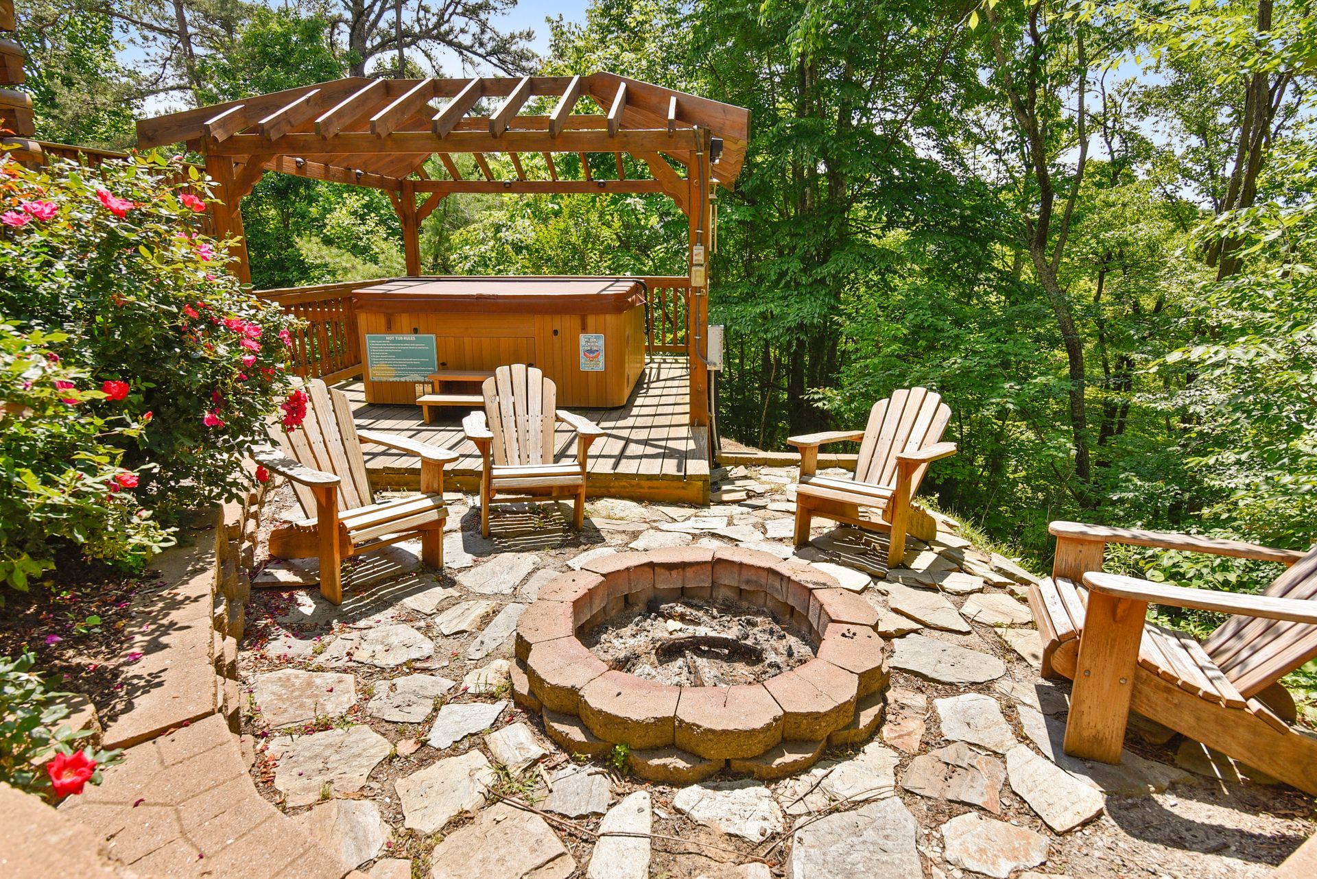 Outdoor patio with hot tub under a pergola, fire pit, and wooden Adirondack chairs, surrounded by greenery.