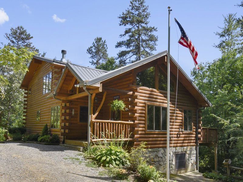 Log cabin with an American flag, surrounded by trees, on a sunny day.