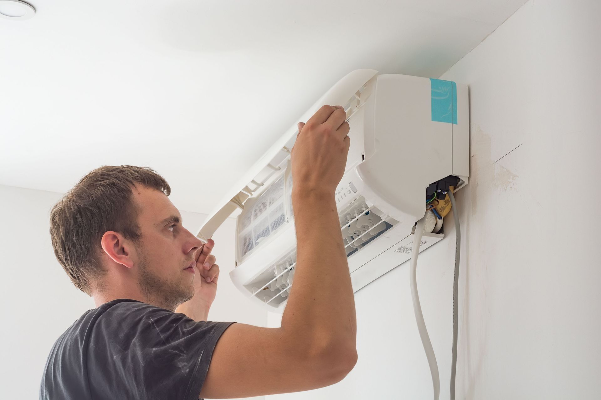 Man installing or repairing a wall-mounted air conditioner unit. White interior, wires visible.