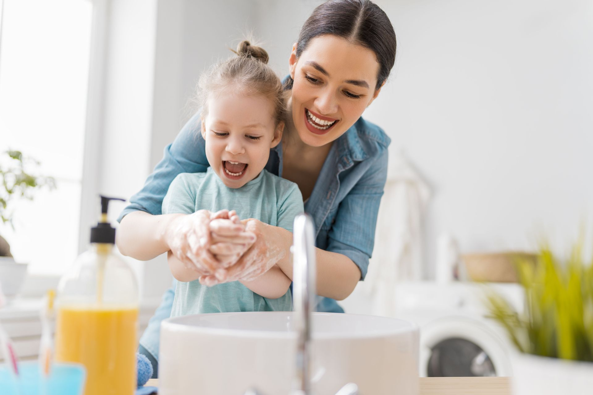 Woman and child washing hands together at a sink, both smiling, in a bright bathroom.