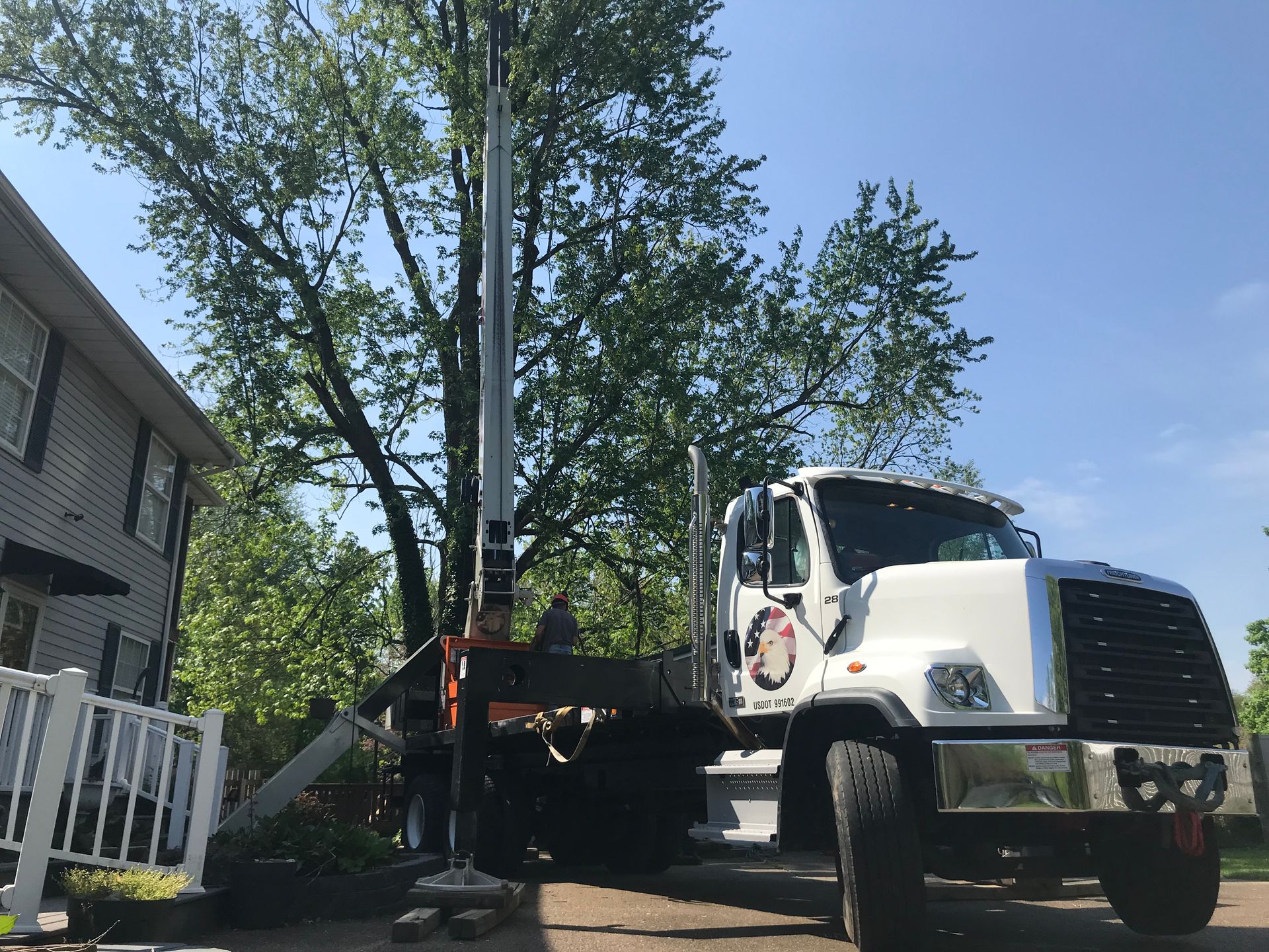 A white boom truck is parked next to a house with its crane extended toward a large, leafy tree.