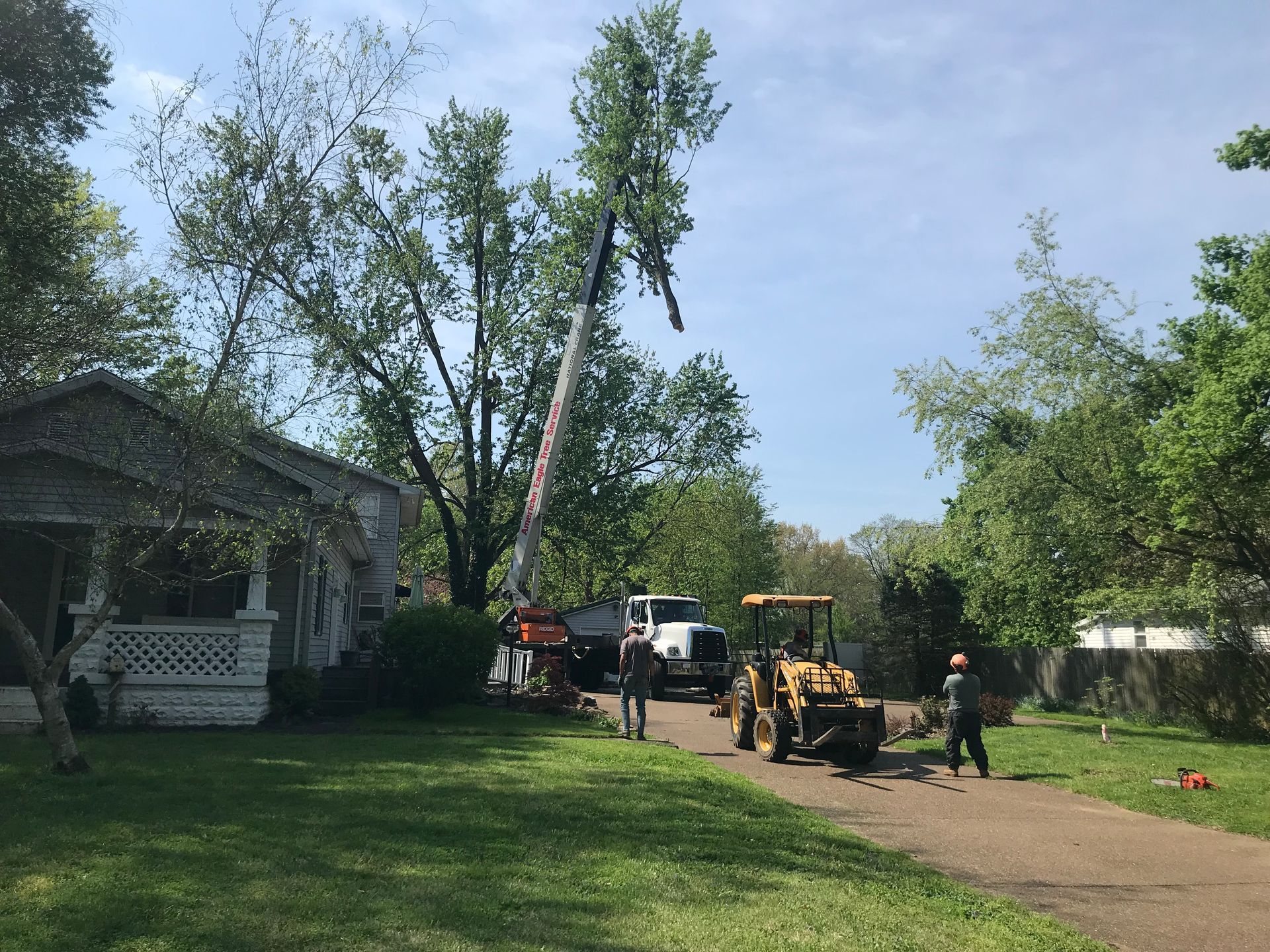 Workers use a crane and heavy machinery to trim a large tree near a house on a sunny day.