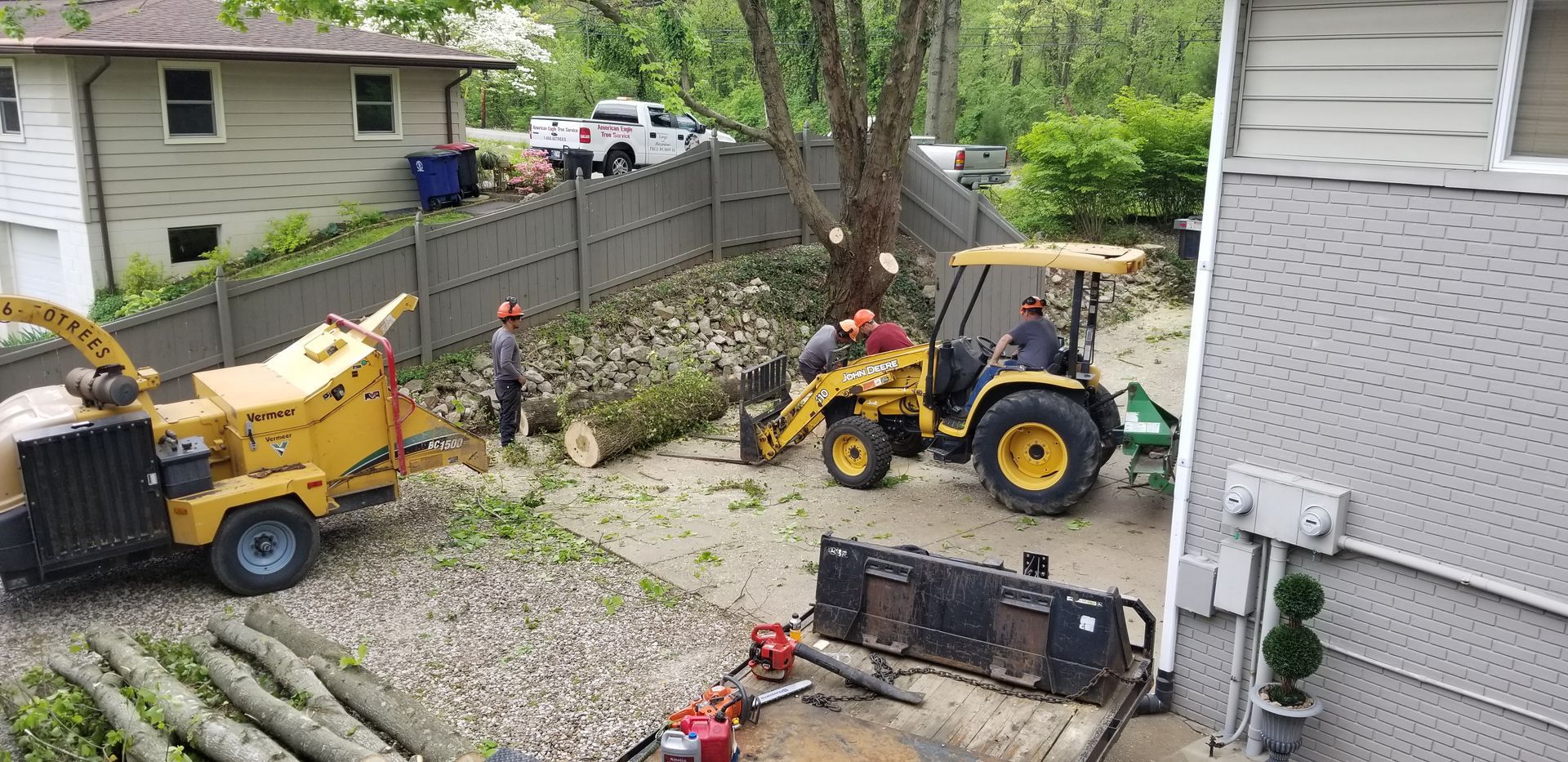 A yellow wood chipper and a front-end loader operate in a residential yard as workers clear tree limbs and logs.