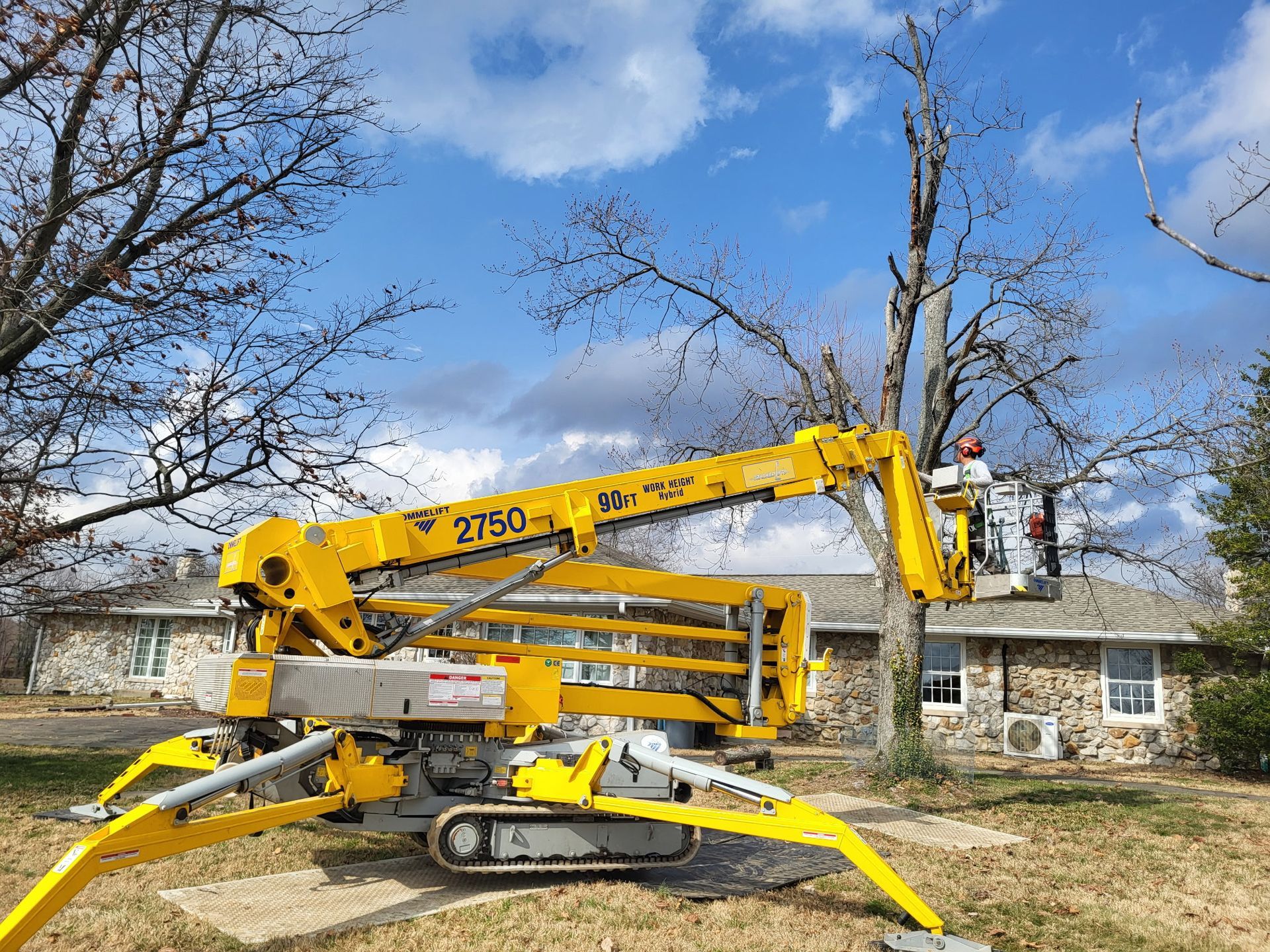 A yellow tracked lift, positioned on a residential lawn, reaches up to trim tree branches near a stone house.