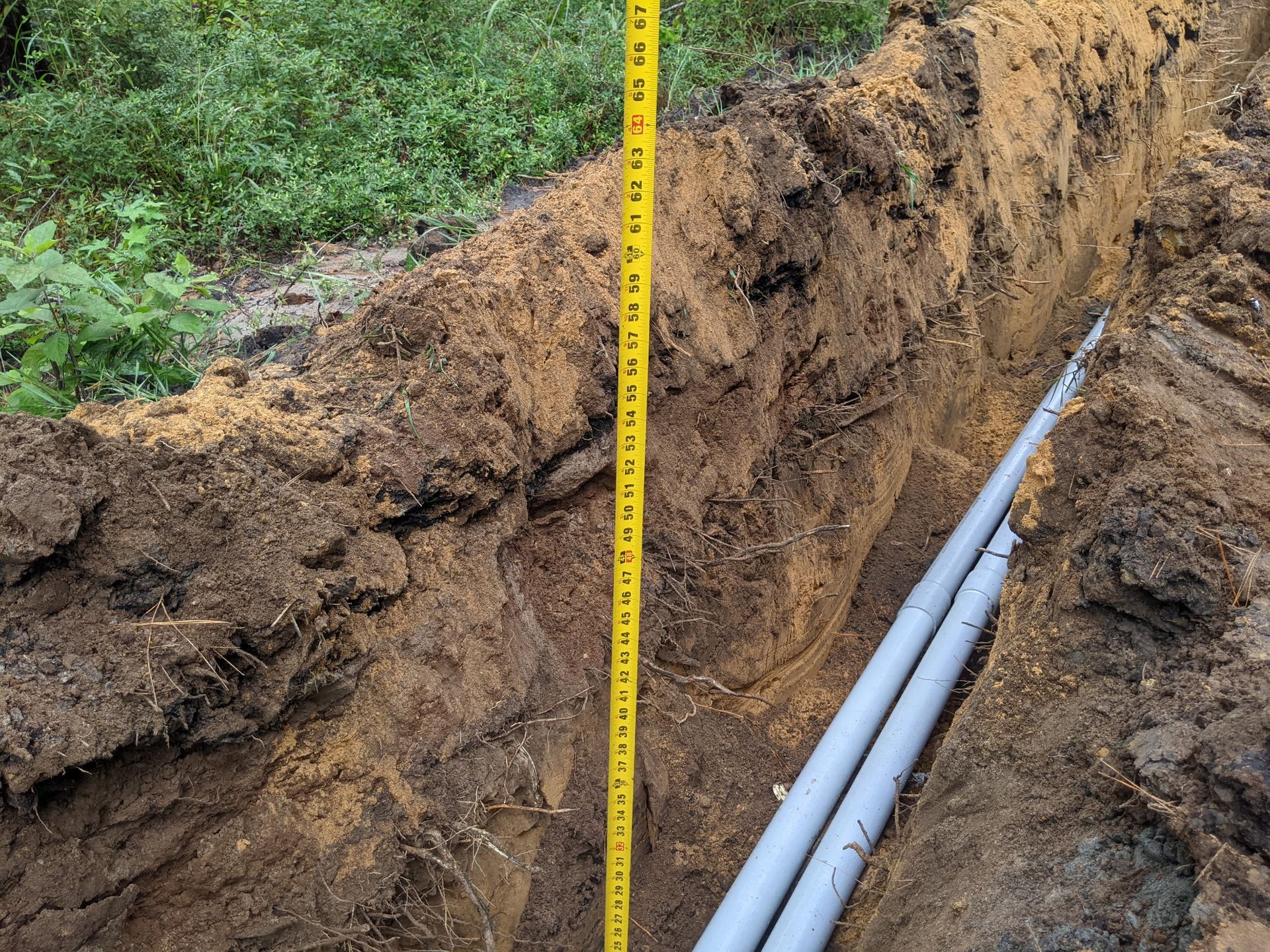 A trench with gray pipes and a yellow tape measure inside; dirt and foliage background.