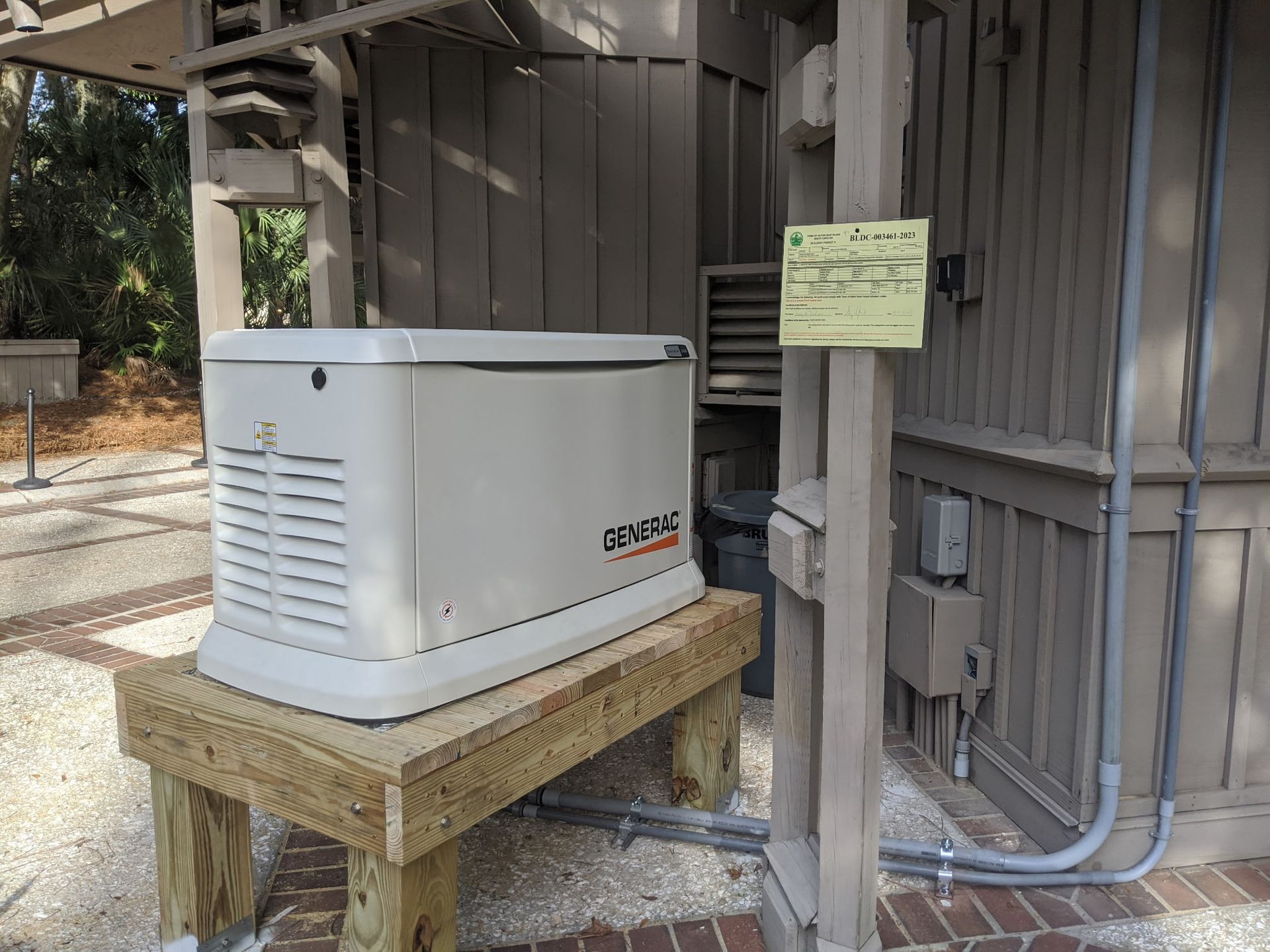 A beige Generac generator on a wooden platform outside a building; gray walls, brick patio.