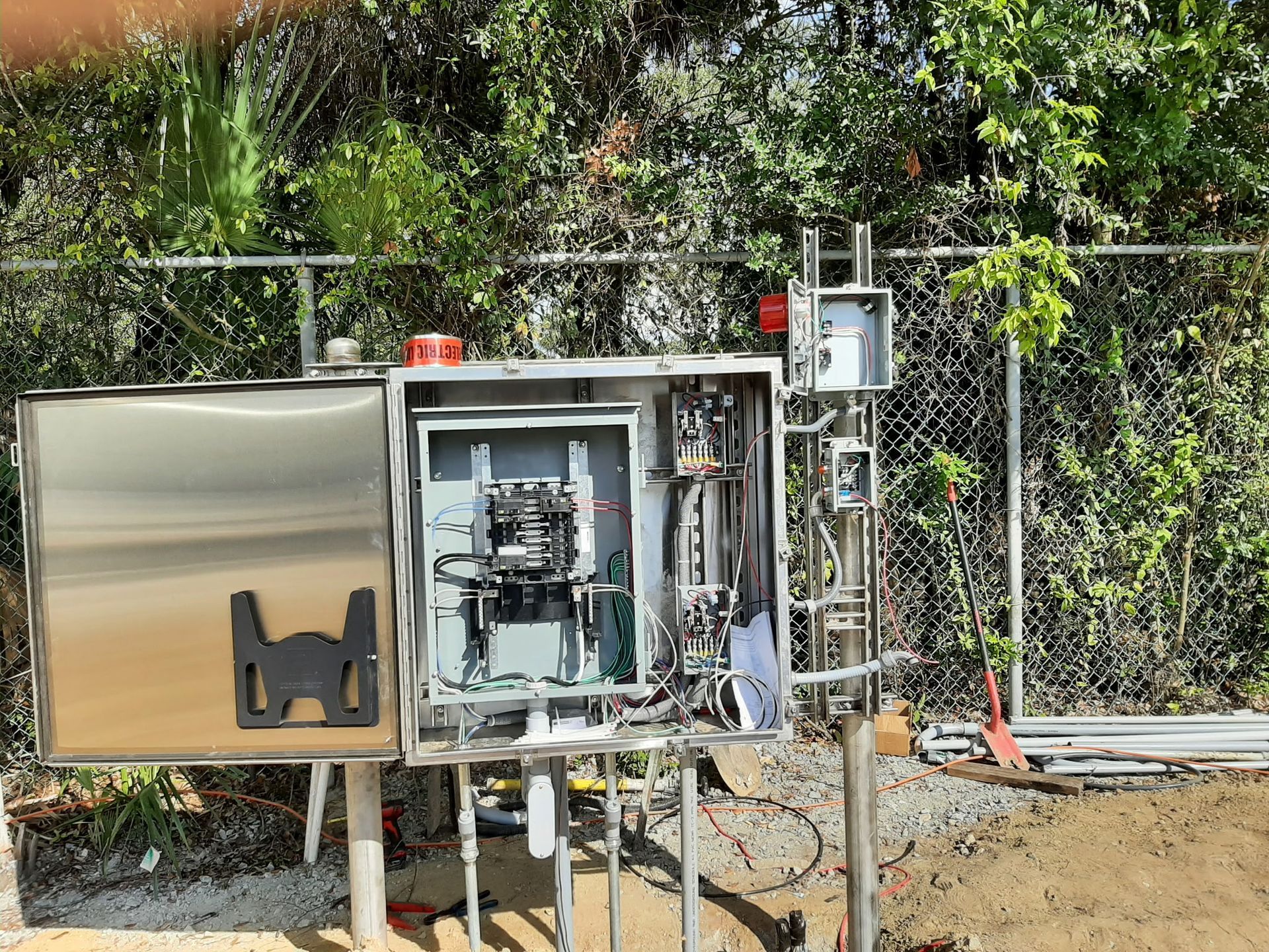 Stainless steel electrical control box outdoors, open to show wires and components, near a fence.