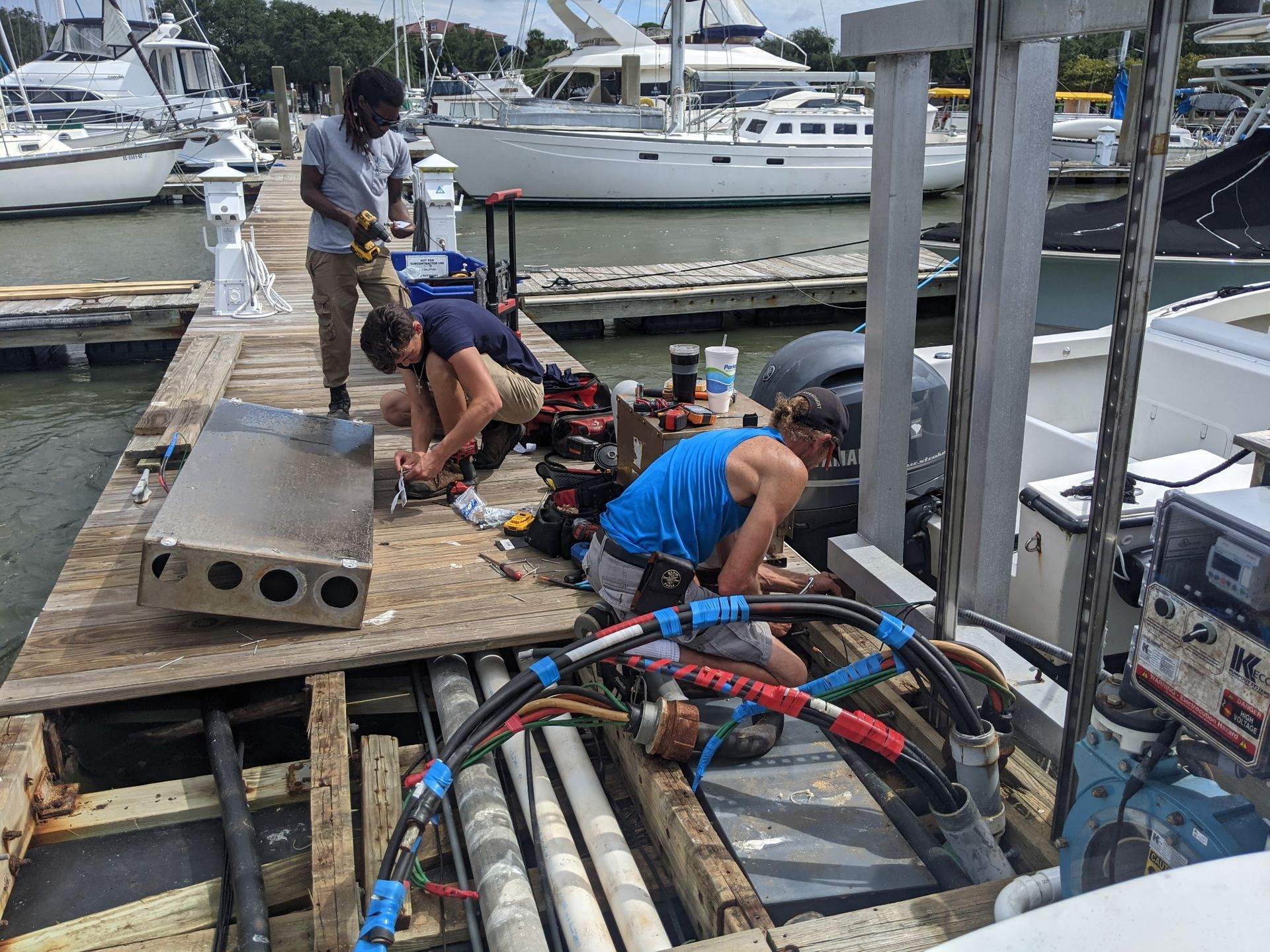 Three people repairing boat machinery on a wooden dock at a marina.