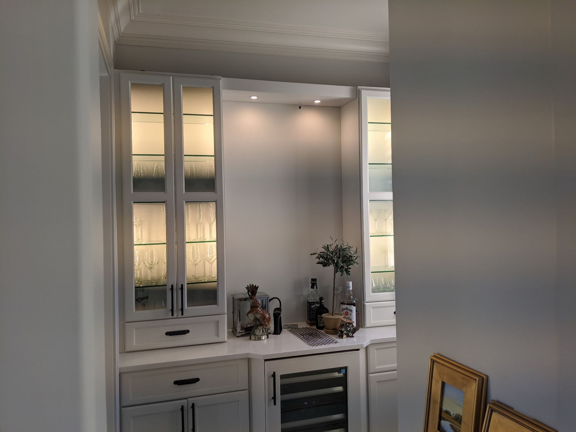 Built-in white bar cabinet with glass-front cabinets, wine fridge, and countertop display with drinks and decorations.
