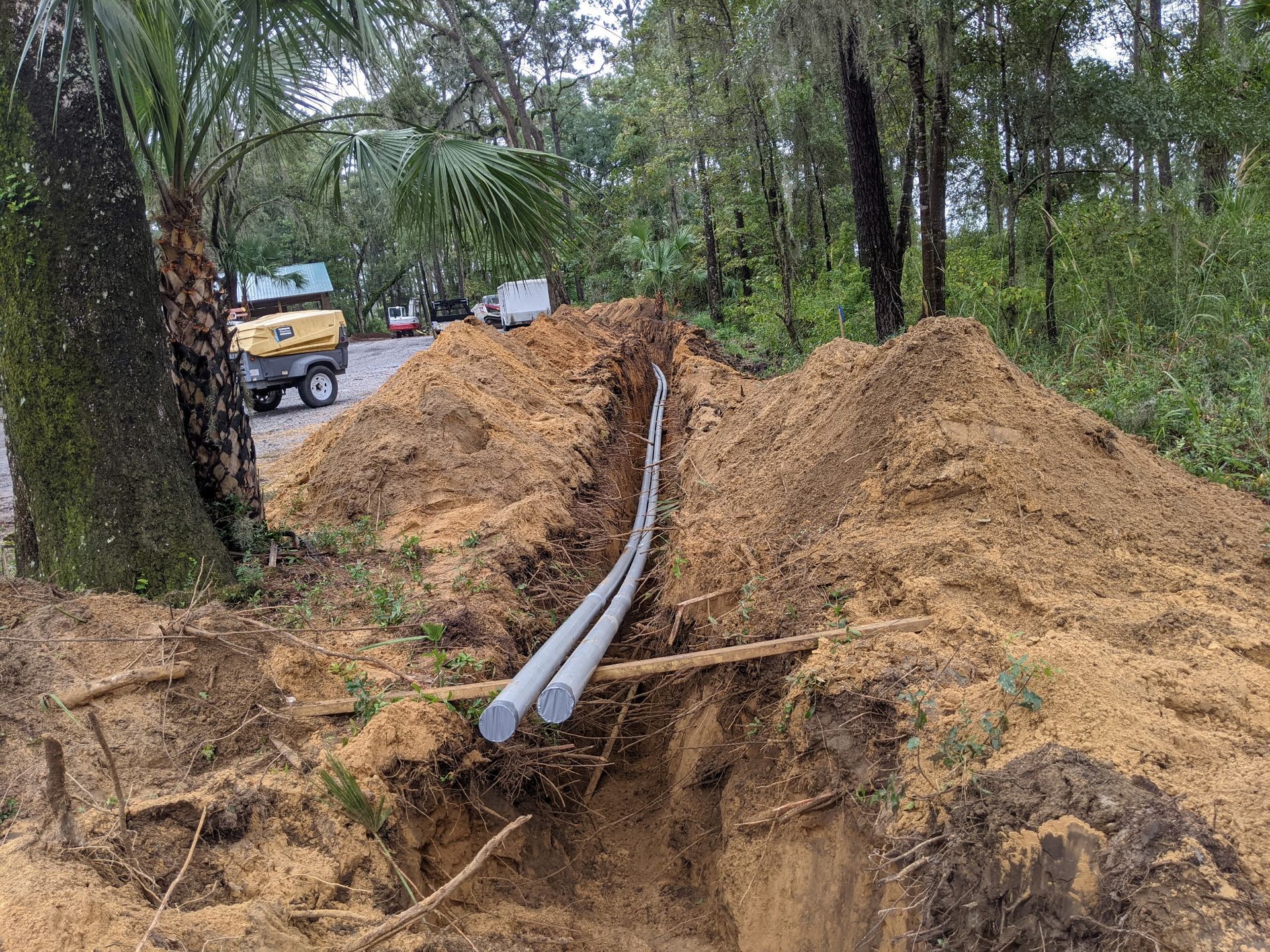 A trench dug in dirt, with gray pipes running through it, and machinery in the background.