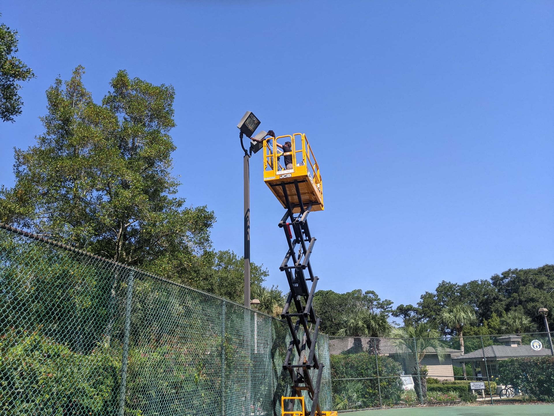 Person in a yellow lift working on a tall black light pole next to a green fence, under a blue sky.