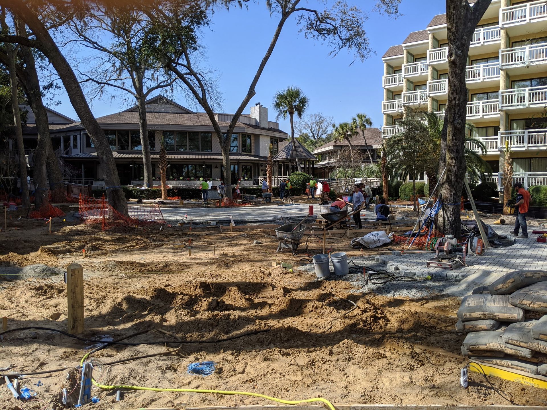 Construction site; dirt ground with workers and equipment in front of buildings and trees under a blue sky.