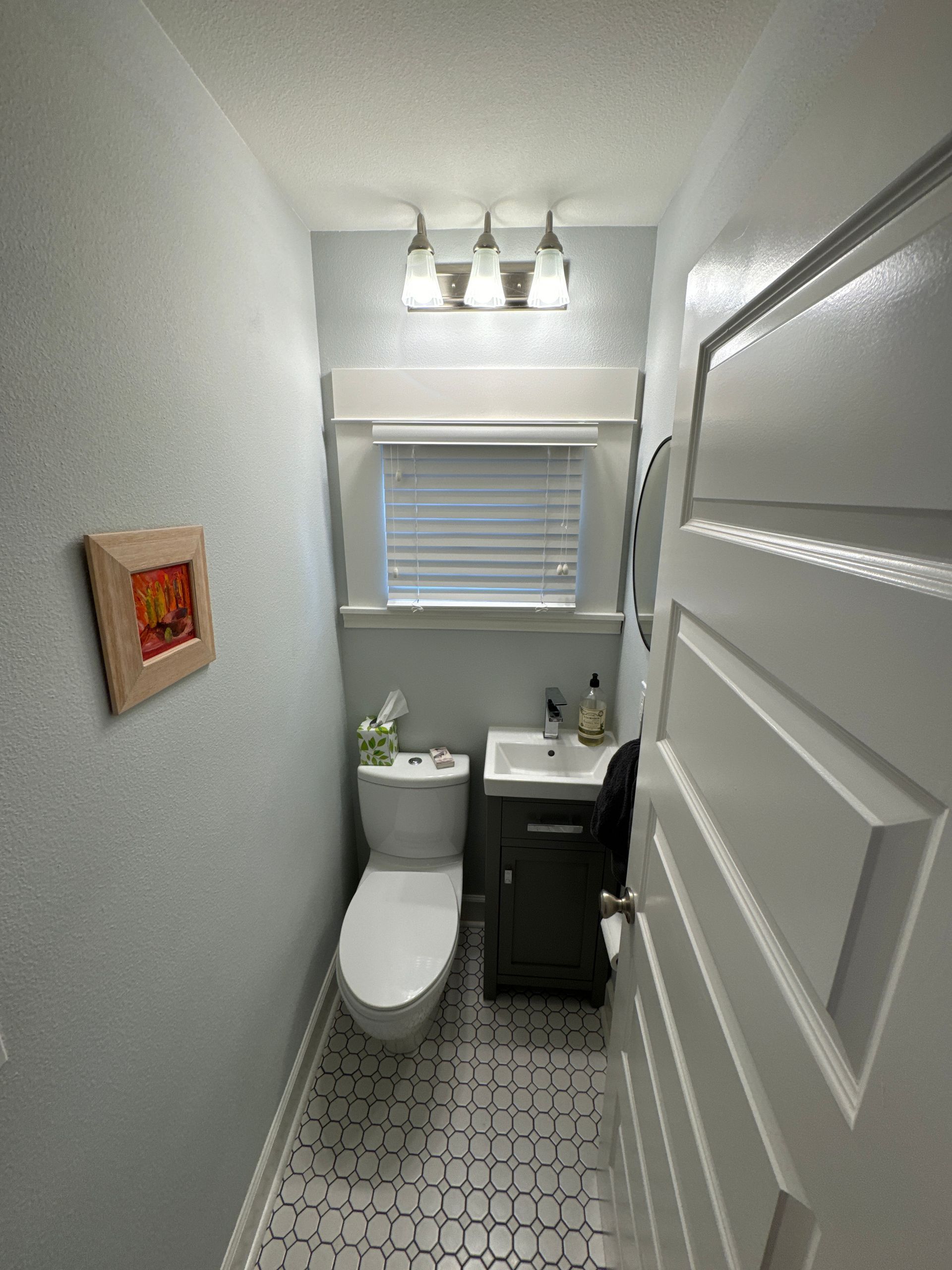 Small narrow bathroom with toilet, sink, window blinds, patterned floor, and overhead lights