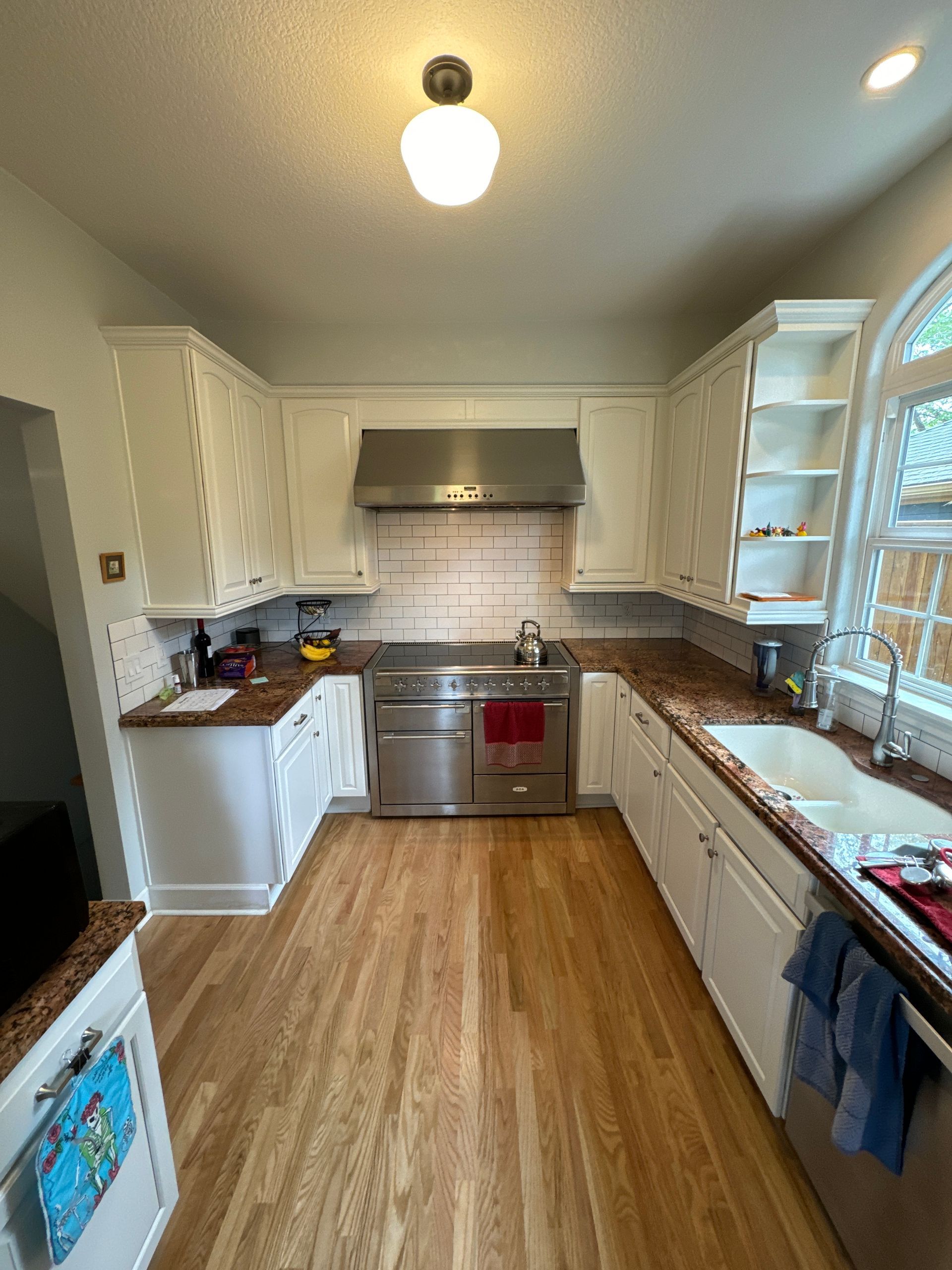 Bright kitchen with white cabinets, stainless steel stove, wood floor, and large window over the sink.