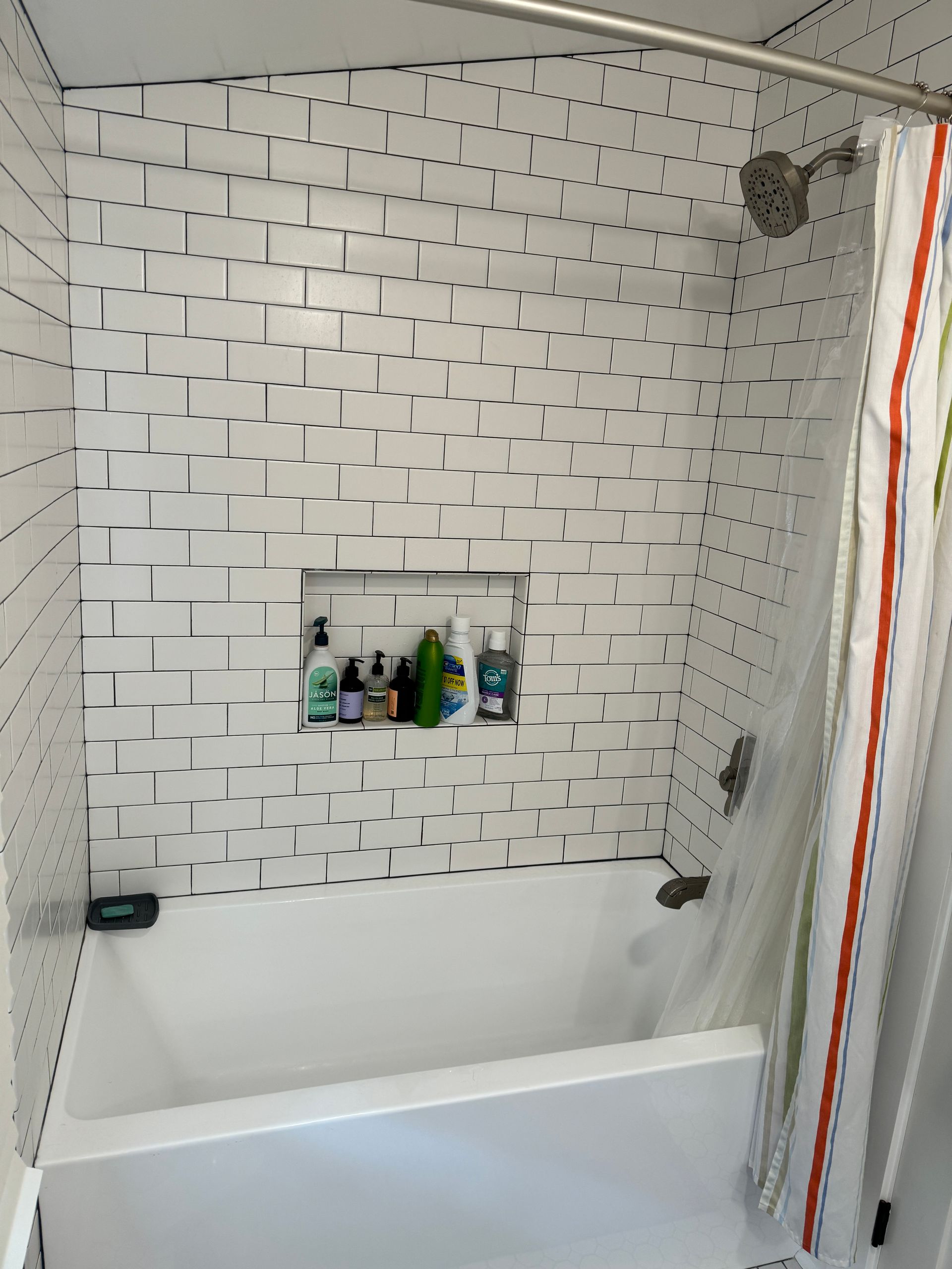 Bathroom shower with white tile, bathtub, and built-in shelf holding toiletries.