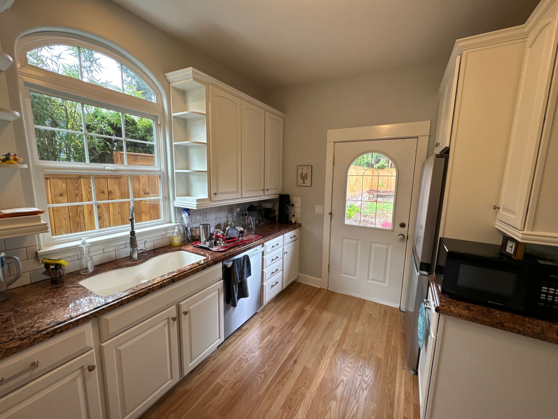 Bright kitchen with white cabinets, wood floors, a large window, and a back door with an arched glass panel