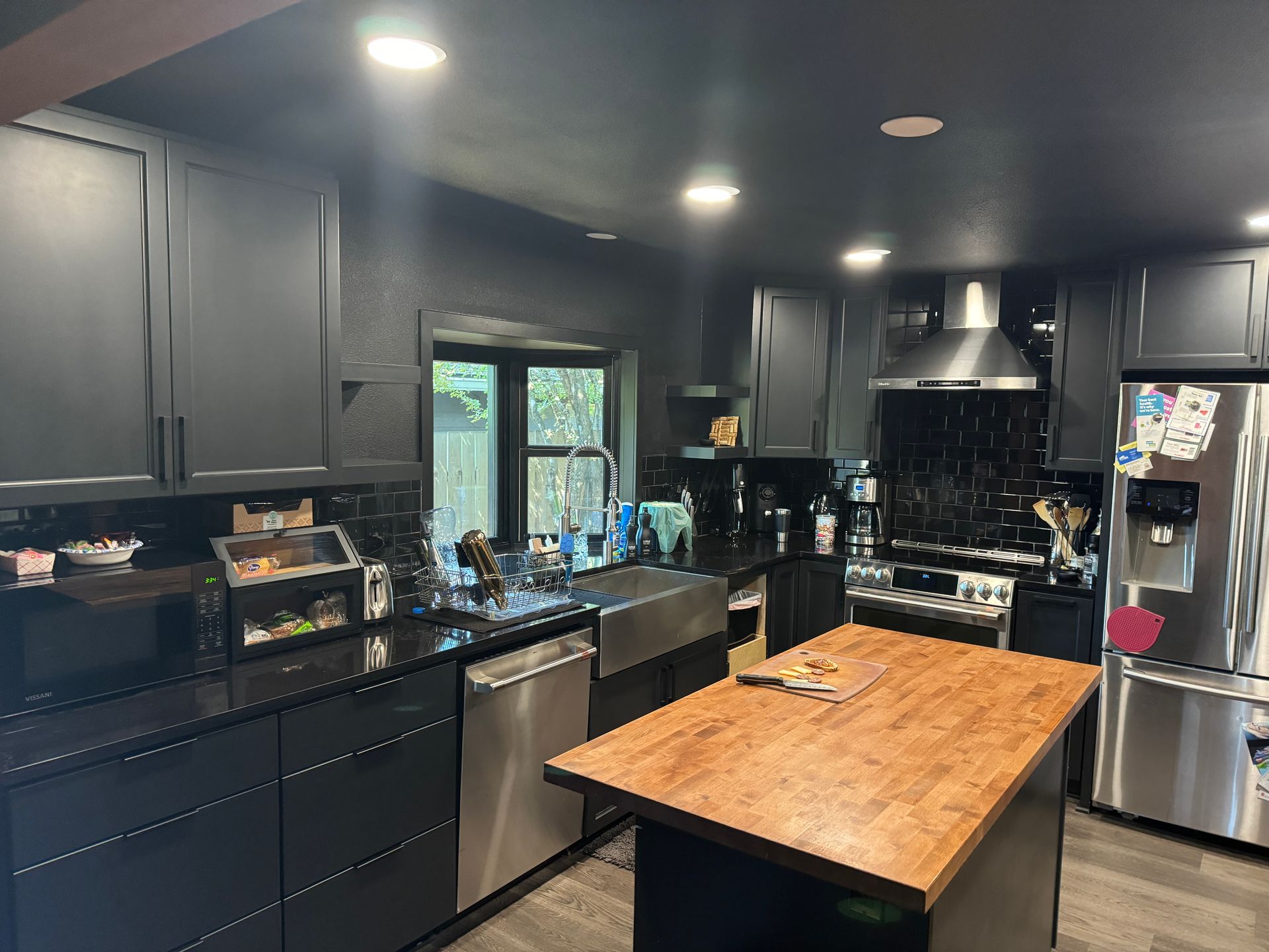 Modern black kitchen with stainless appliances, dark cabinets, and a wooden island under recessed lights
