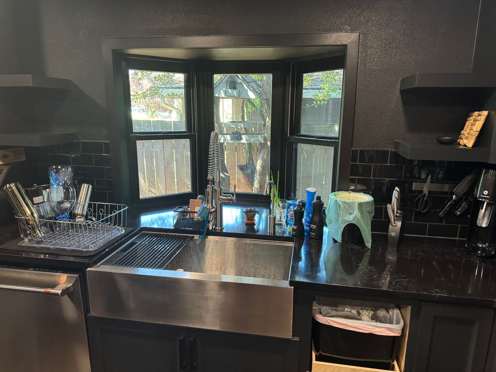 Black kitchen sink beneath a window, with dishes, drying rack, and countertops cluttered with items