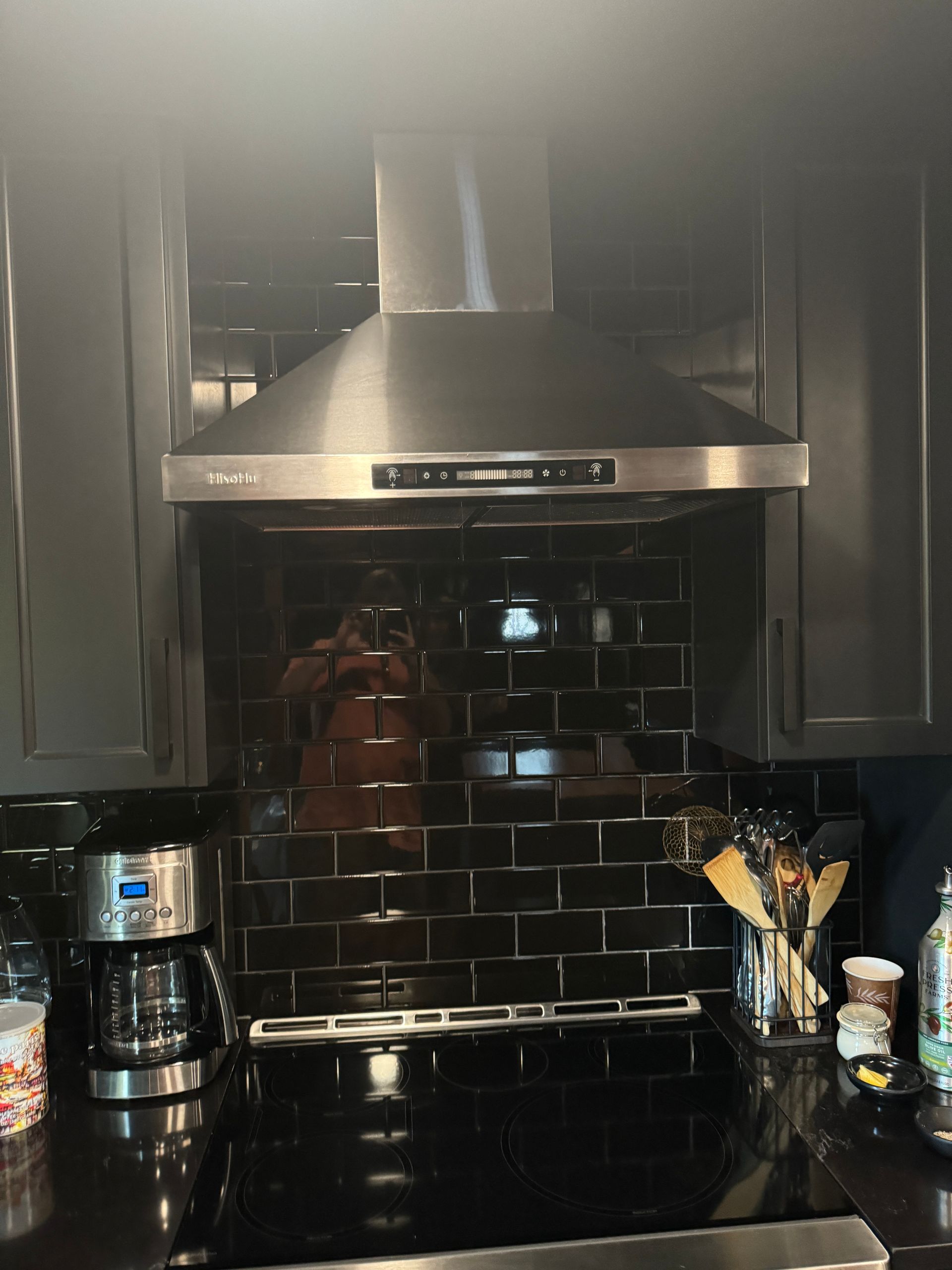 Kitchen stove with stainless steel range hood, black tile backsplash, and countertop items on both sides
