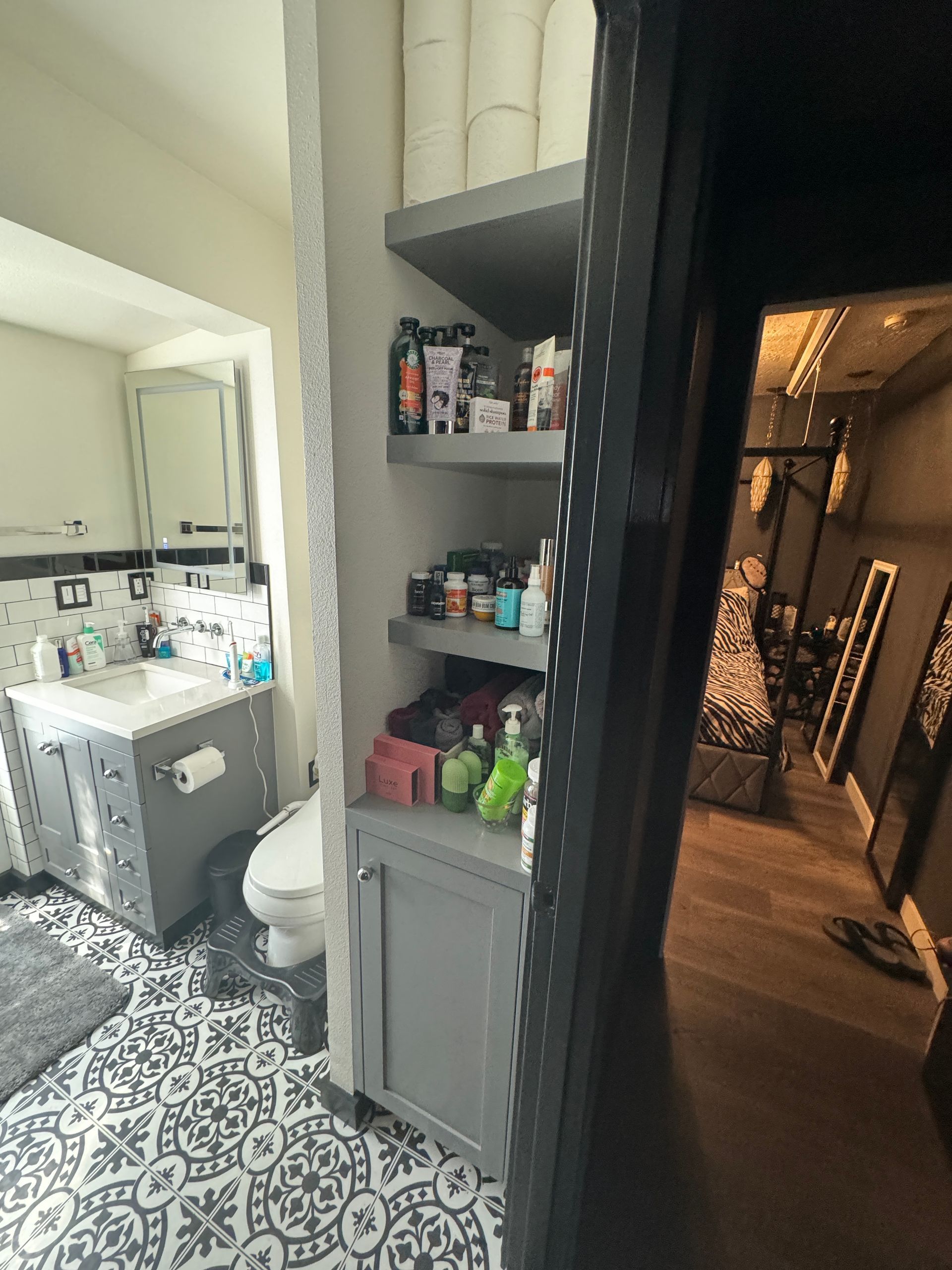 Bathroom with patterned tile, narrow shelf of toiletries, and a dark hallway beyond