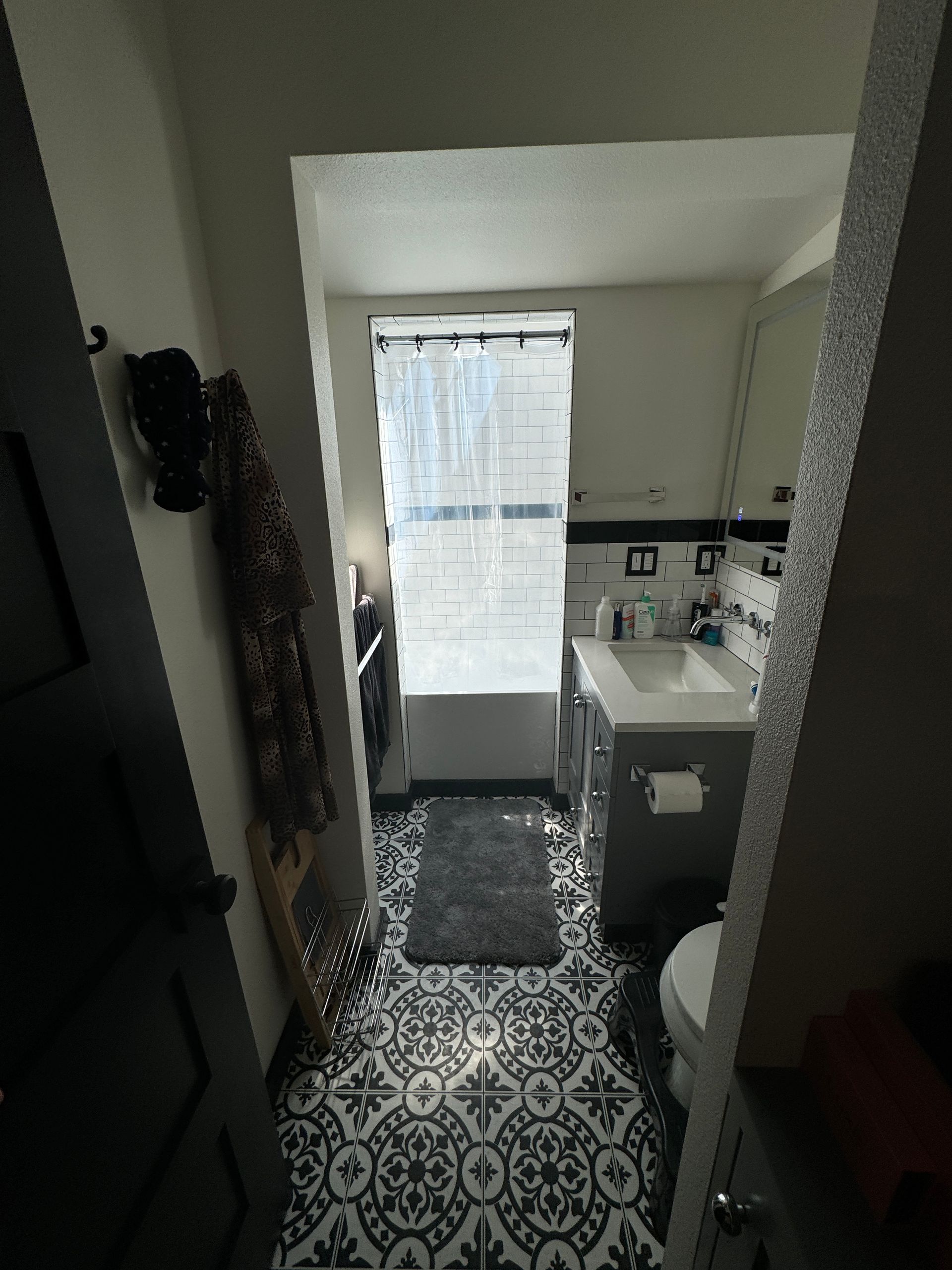 Narrow bathroom with black-and-white patterned floor, vanity sink, and bright window with sheer curtain.