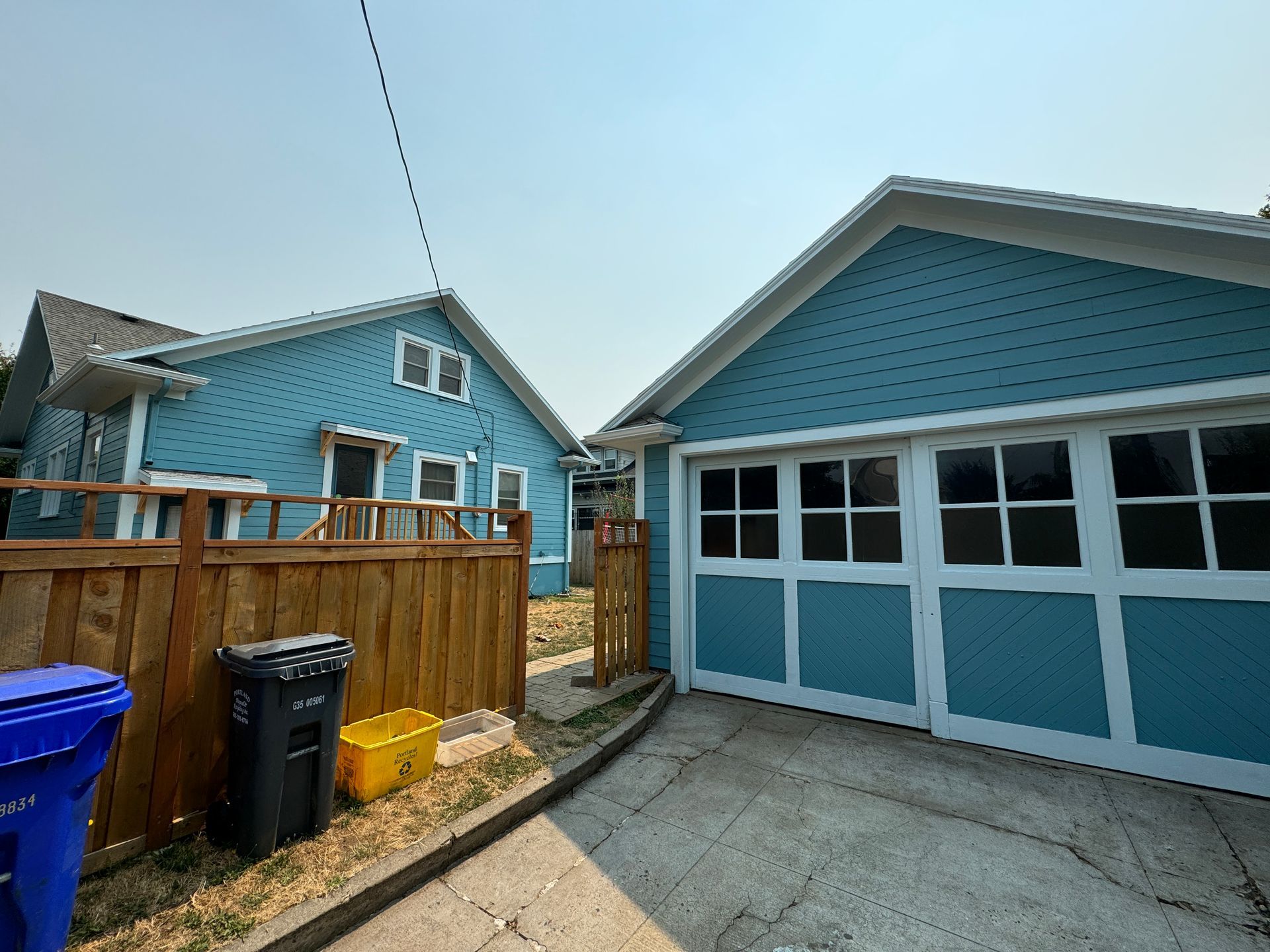 Blue house and garage with fenced yard, trash bins, and paved driveway under a clear sky