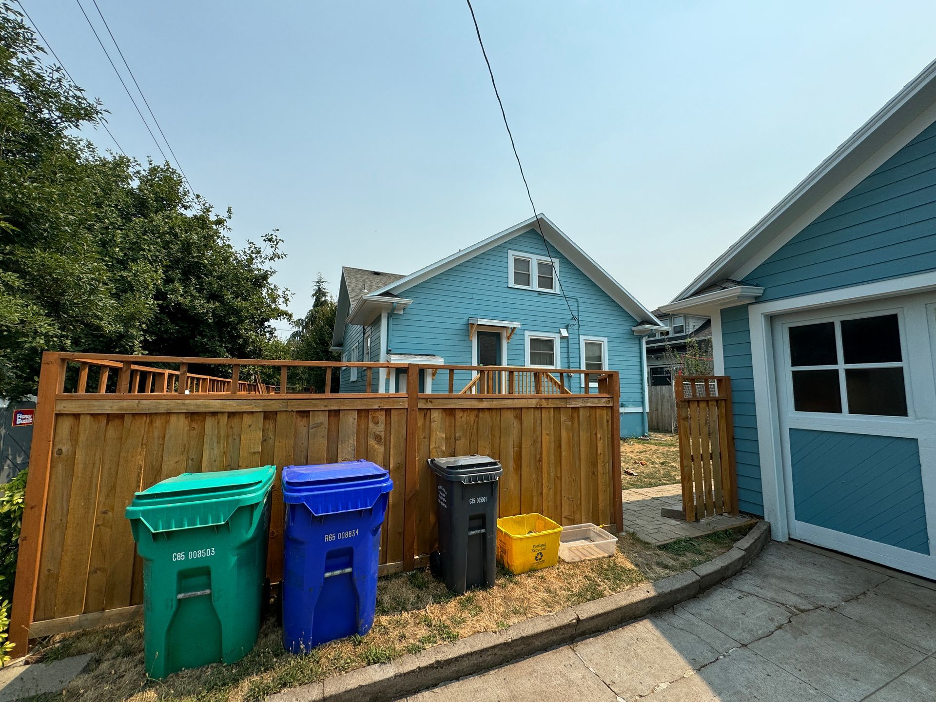 Backyard beside blue house with fenced trash bins and driveway to garage