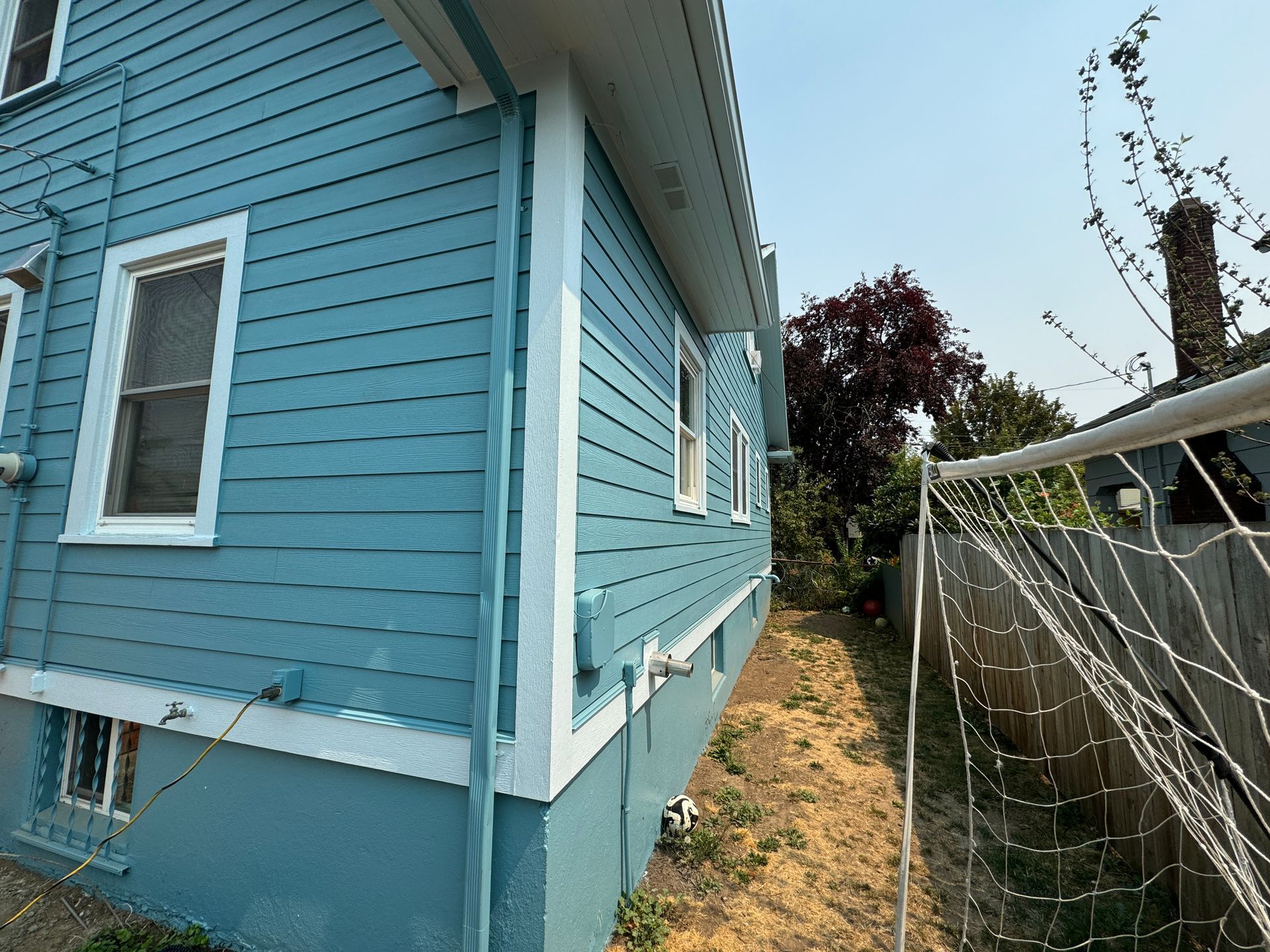 Blue house side yard with narrow dirt path beside a chain-link fence