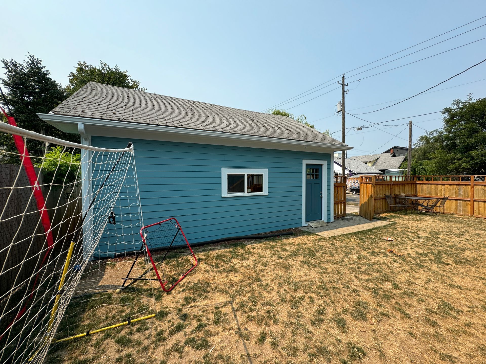 Blue shed in a backyard with a white roof, fence, and swing set on dry grass.