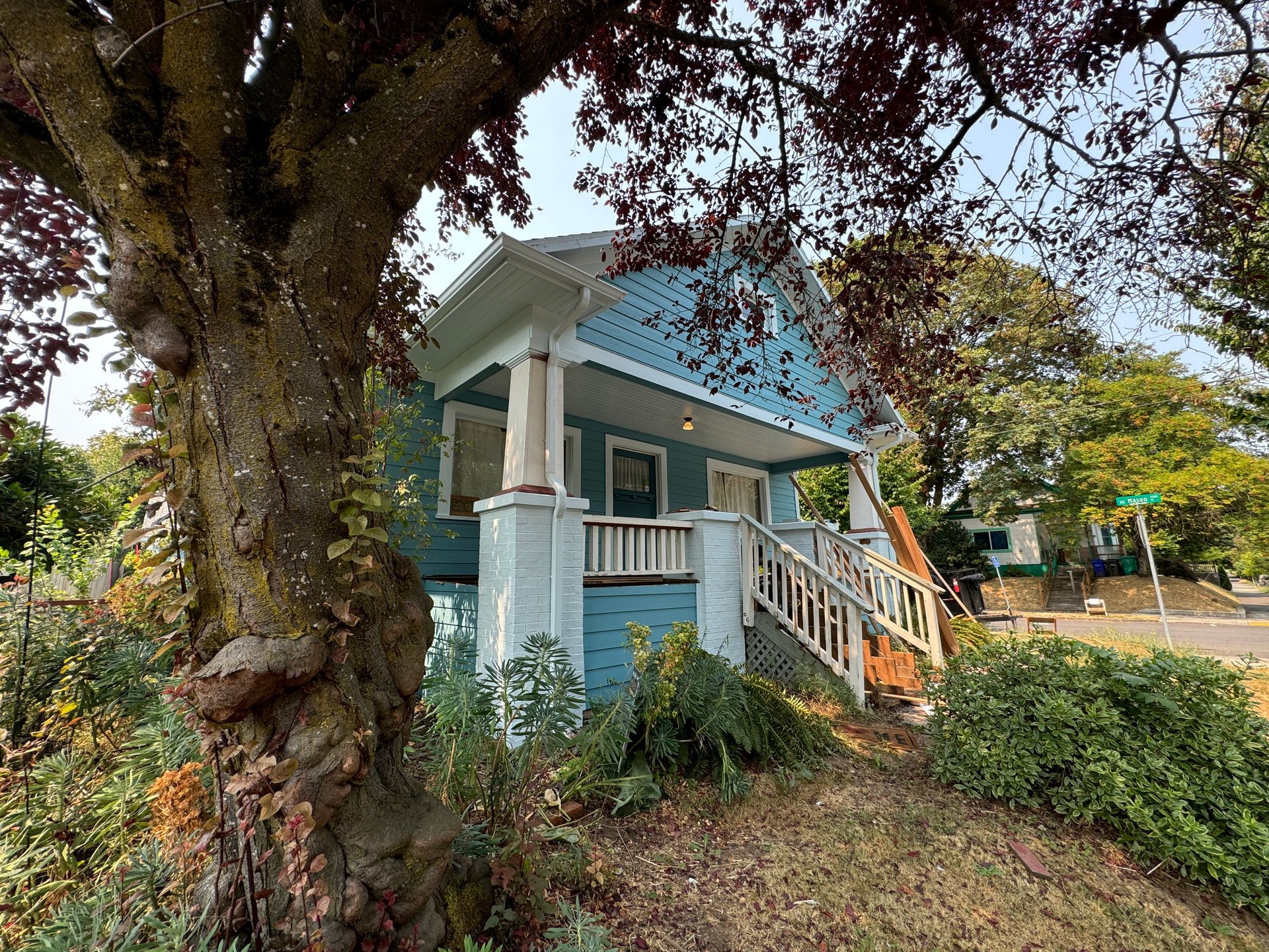Blue Victorian-style house with white trim and porch, partly shaded by large trees and surrounded by greenery