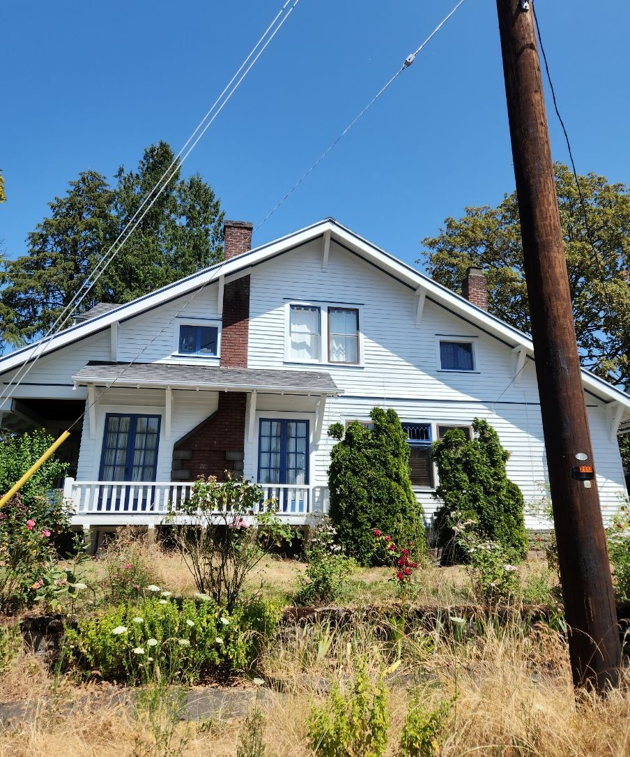 White house with a front porch, green shrubs, and a wooden utility pole under a clear blue sky