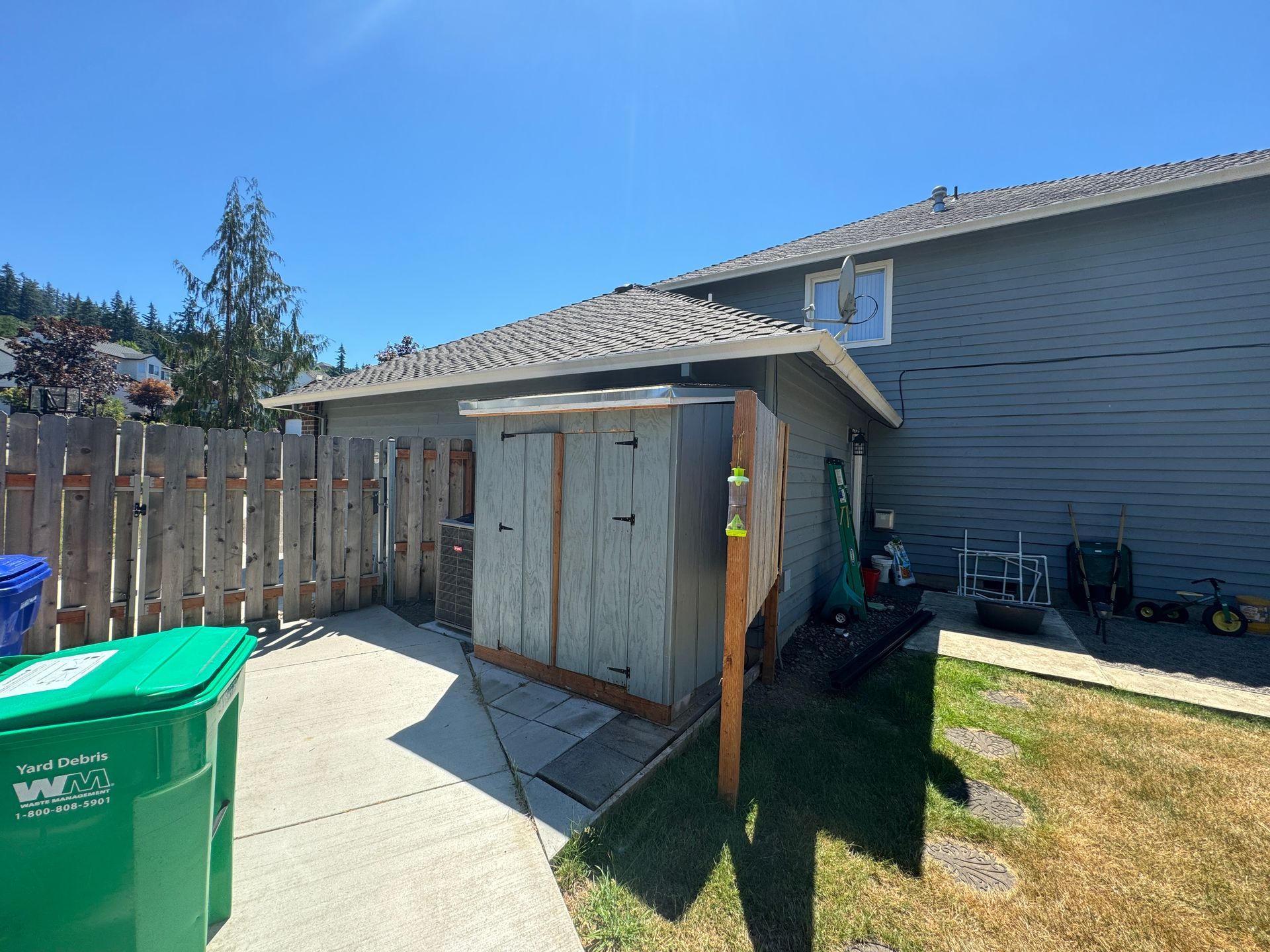 Backyard shed beside a house, with wooden fence, trash bin, and sunny blue sky.