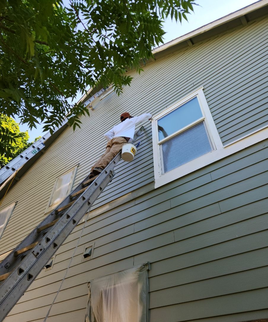 Worker on a ladder painting the exterior siding of a gray house near a window.