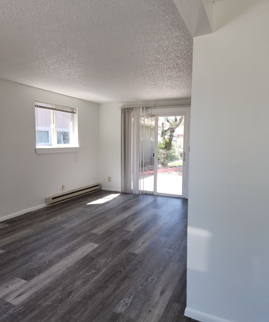 Empty living room with gray wood floors, white walls, a window, and sliding glass doors to a patio.