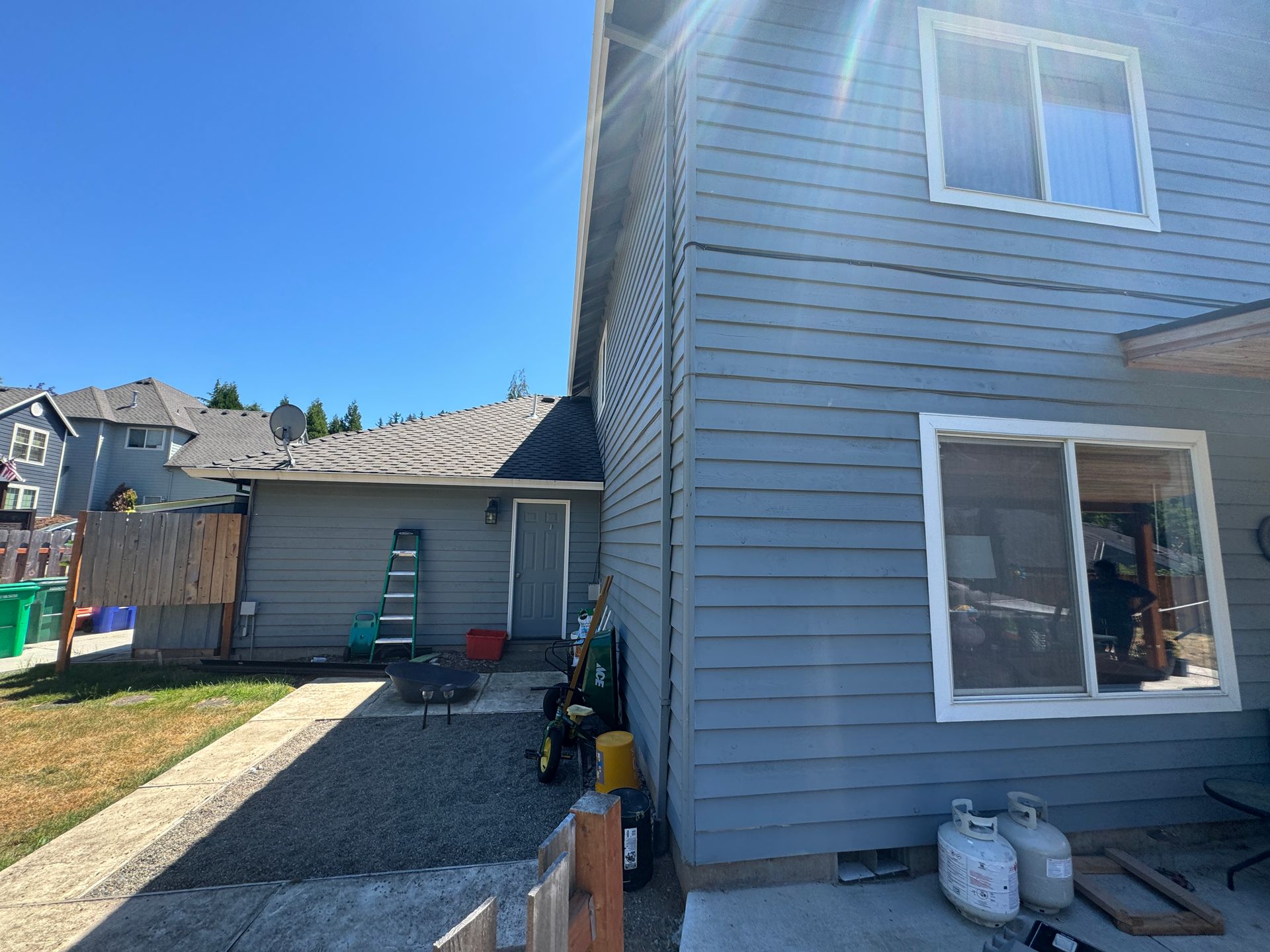 Backyard view of gray house siding, driveway, shed, and patio furniture under a clear blue sky