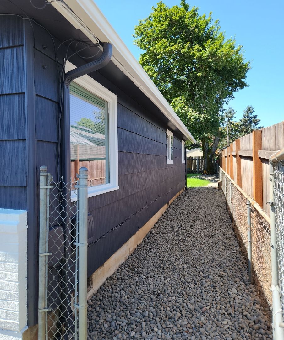 Narrow gravel path between a dark blue house and wooden fence, with a tree at the end.