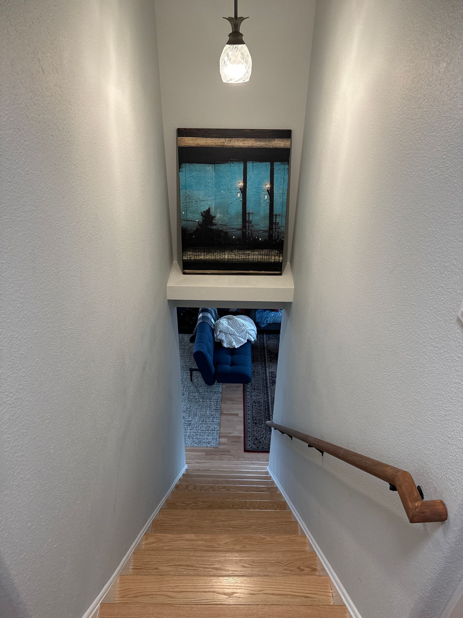 Narrow stairway with wooden steps, wall art, and a lit ceiling pendant leading to a blue room below