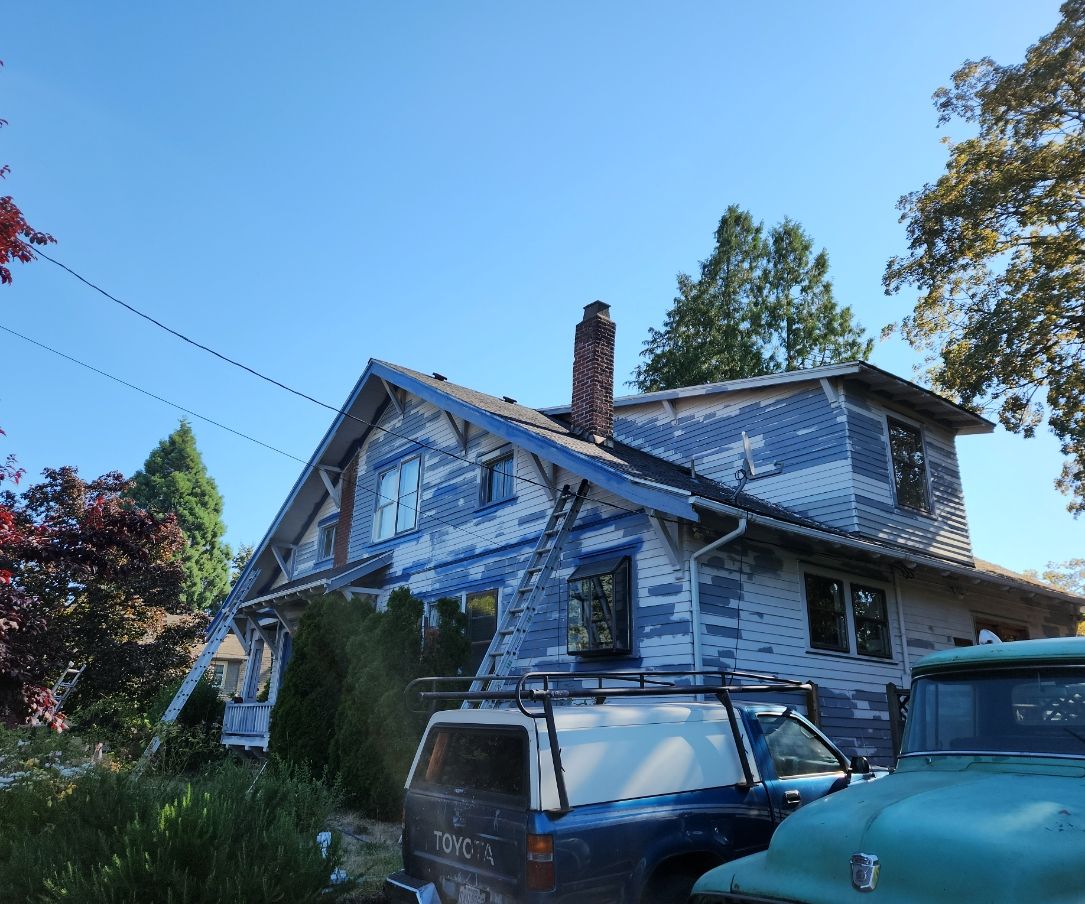 Weathered blue house with steep roof, trees around it, and two parked vehicles in front