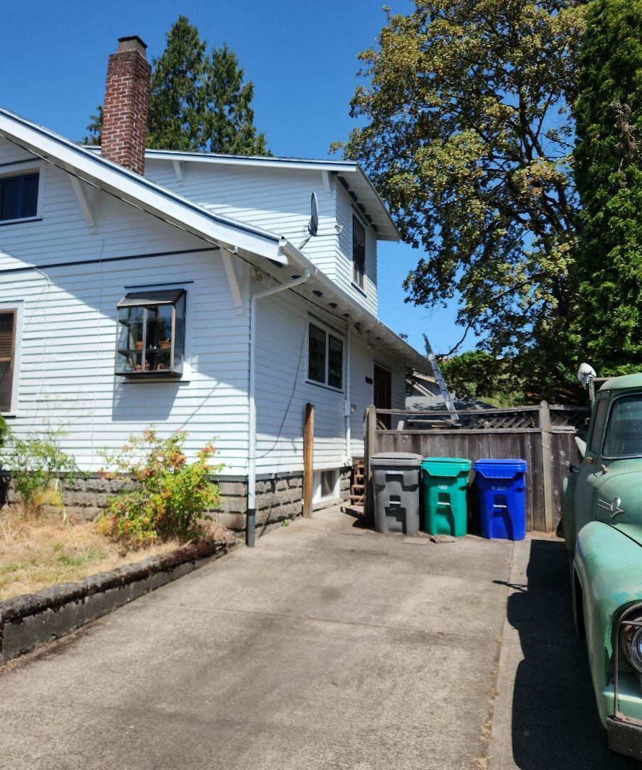 White house with driveway, trash bins, and a green vehicle parked beside it on a sunny day