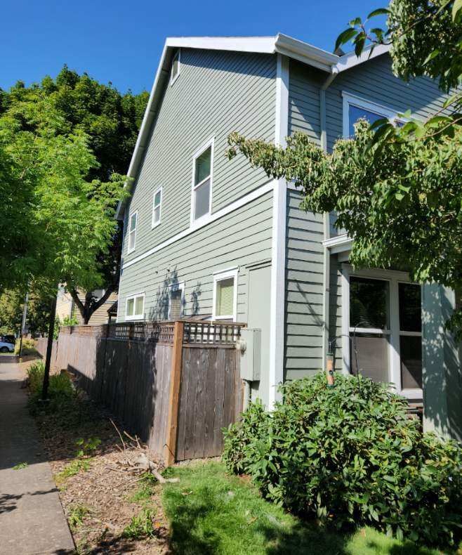 Gray two-story house beside a narrow sidewalk, with a wooden fence, bushes, and leafy trees in bright daylight