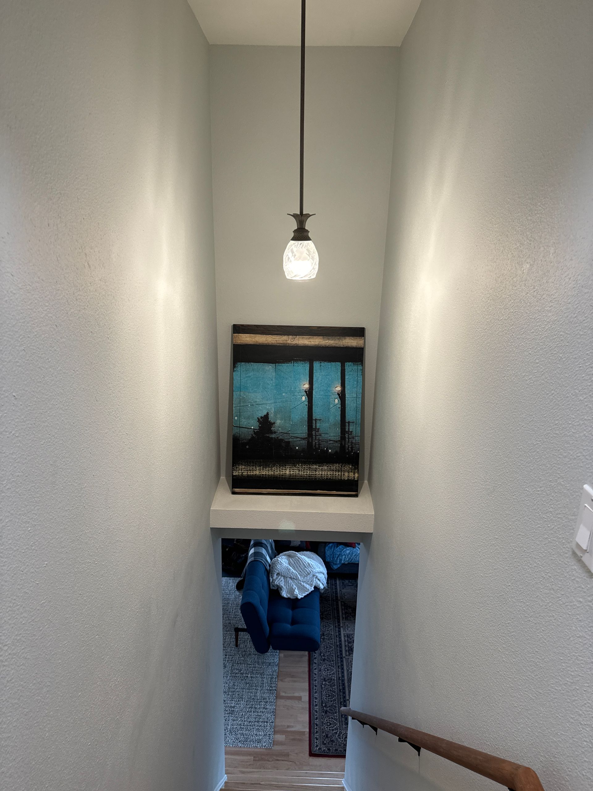 Narrow stairwell with hanging light, framed beach art, and a view into a room below