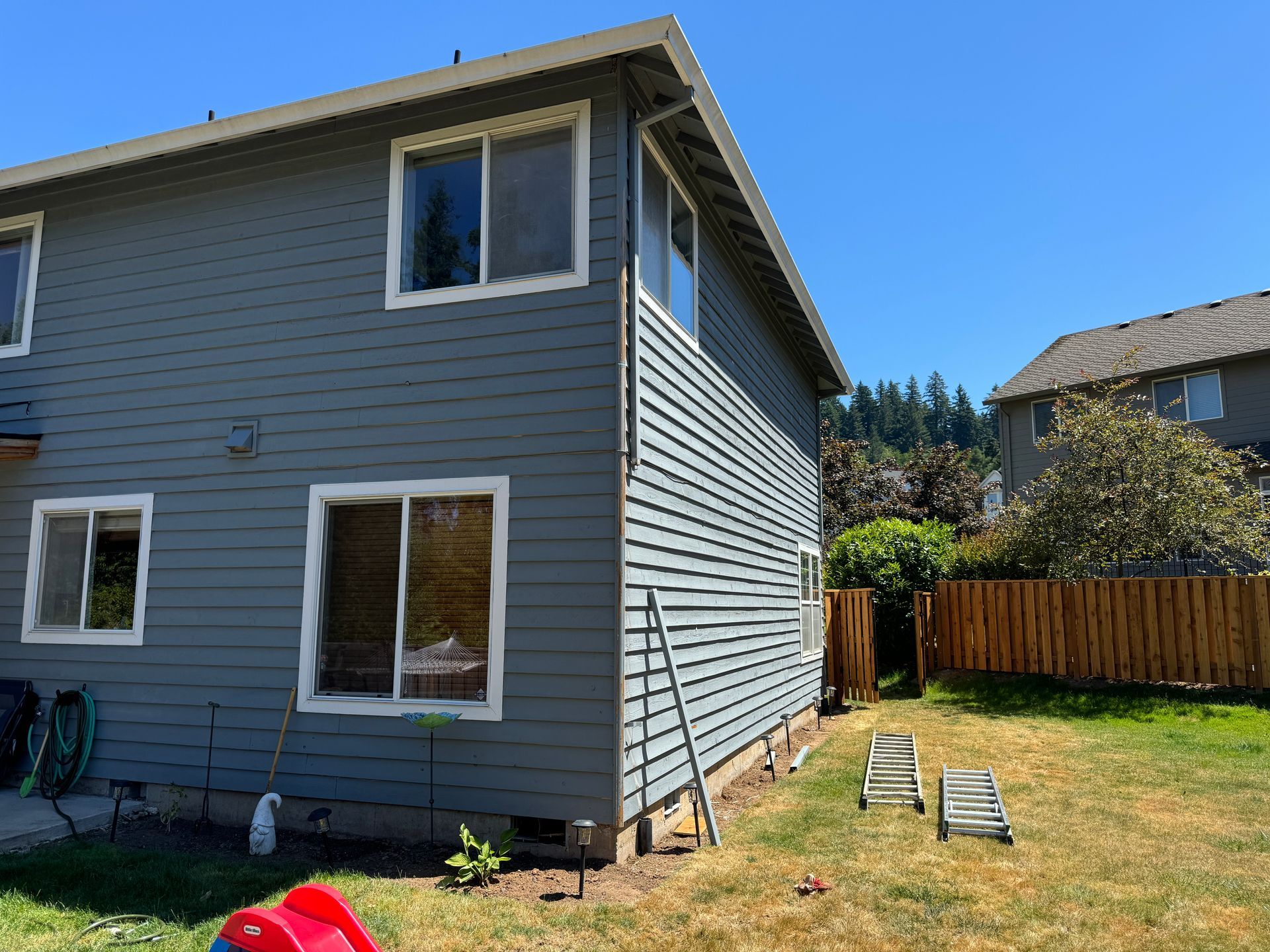 Gray two-story house with white-trim windows and a fenced backyard under a clear blue sky