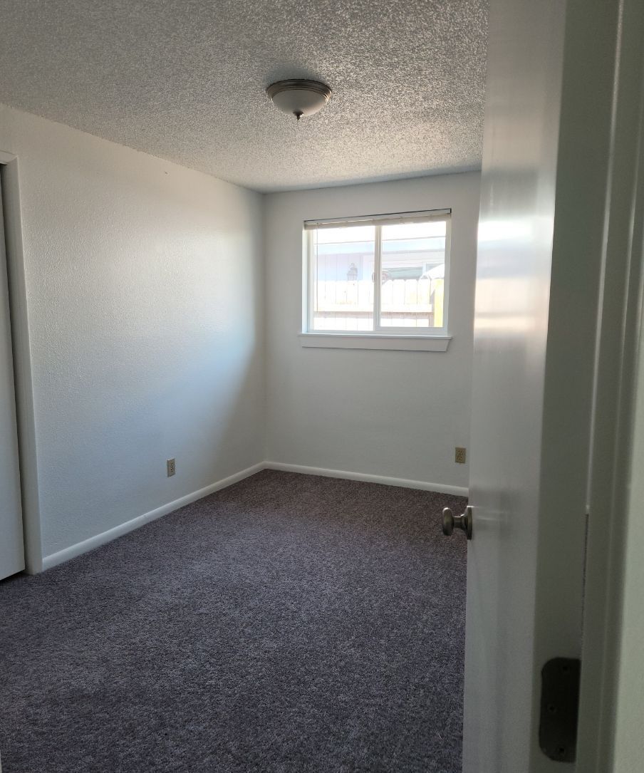 Small empty white bedroom with gray carpet and a window at the far end.