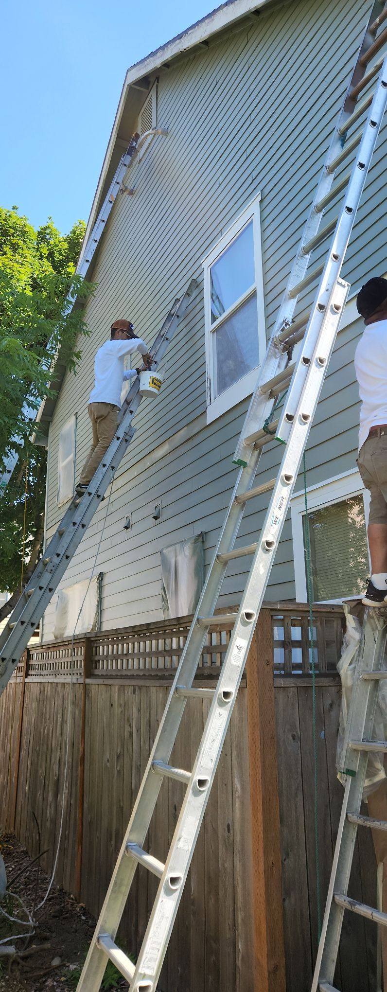 Two people on ladders repairing beige house siding beside a window and wooden fence