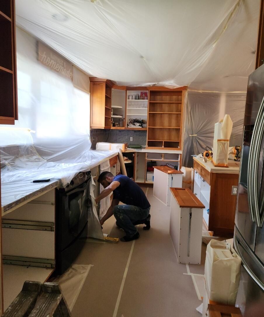Person kneeling and working under a counter in a narrow kitchen room with shelves and covered surfaces.