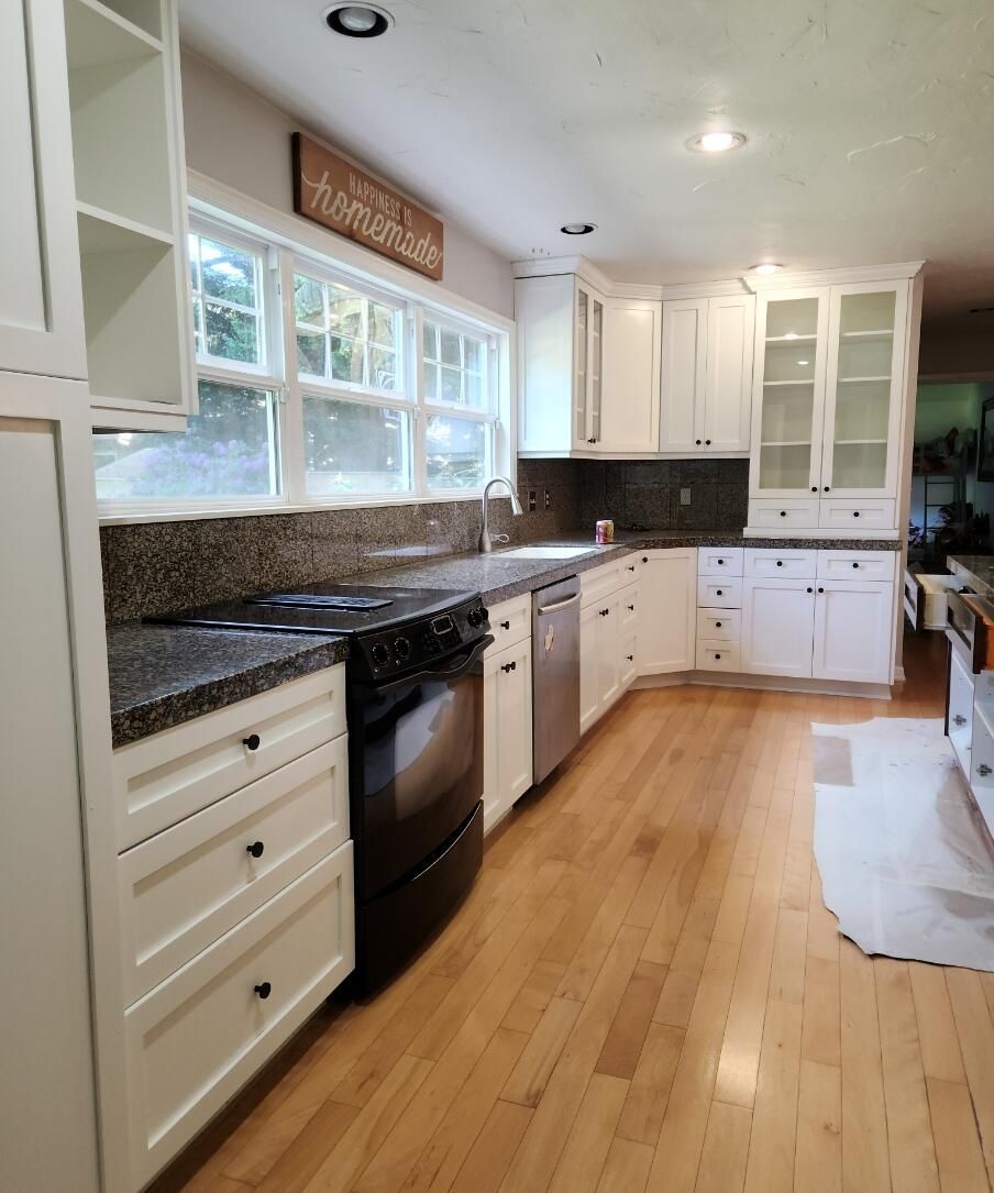 Bright white kitchen with granite counters, black appliances, wood floor, and large windows.