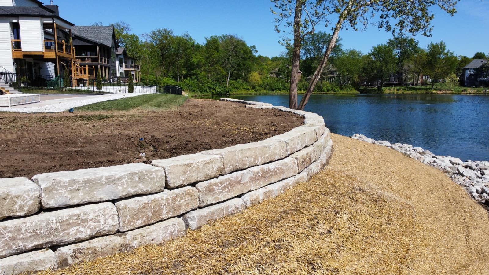 A stone wall is being built next to a lake with a house in the background.