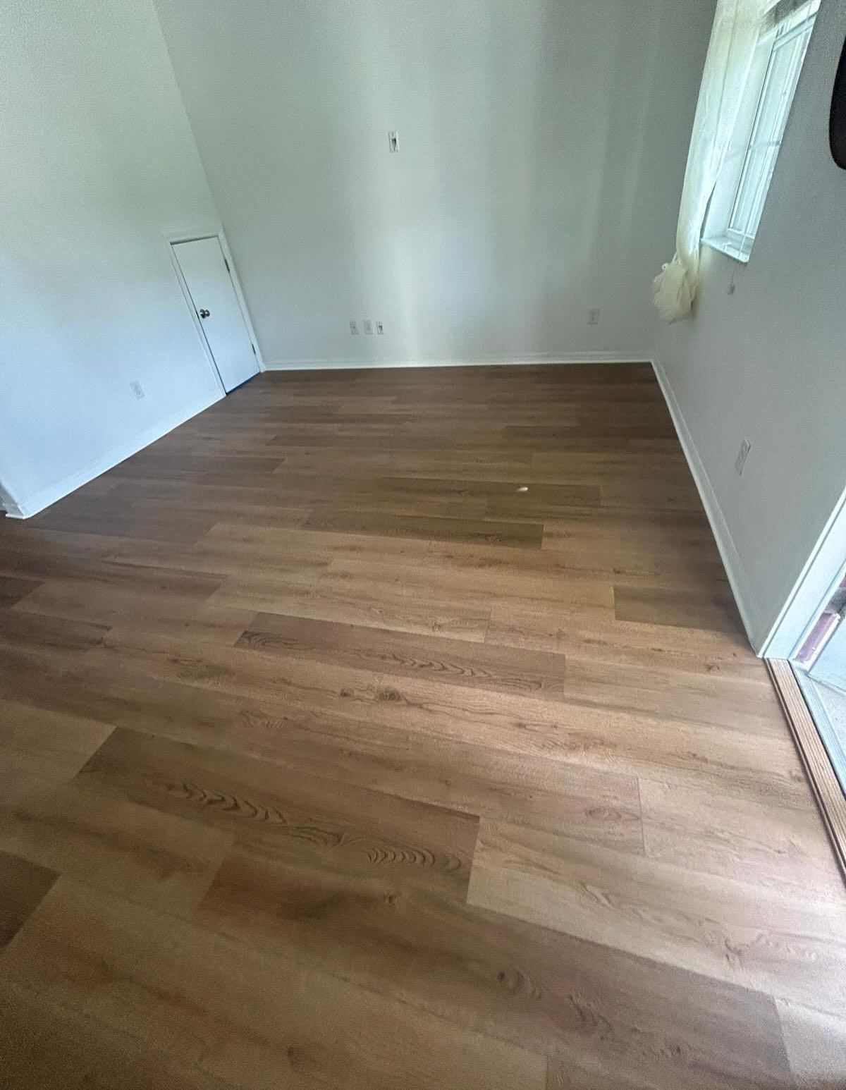 Empty room with brown wood-look flooring and white walls. A small door and a window are visible.