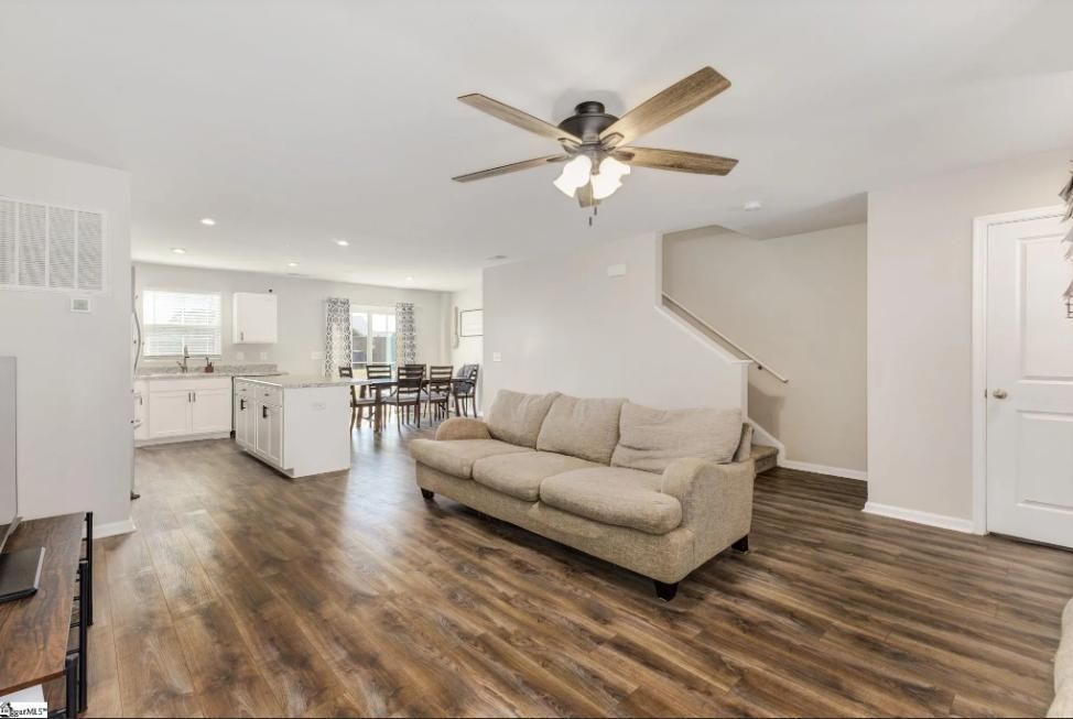 Open-concept living space with kitchen, dining area, and stairs. Beige sofa on wood floors, white walls.